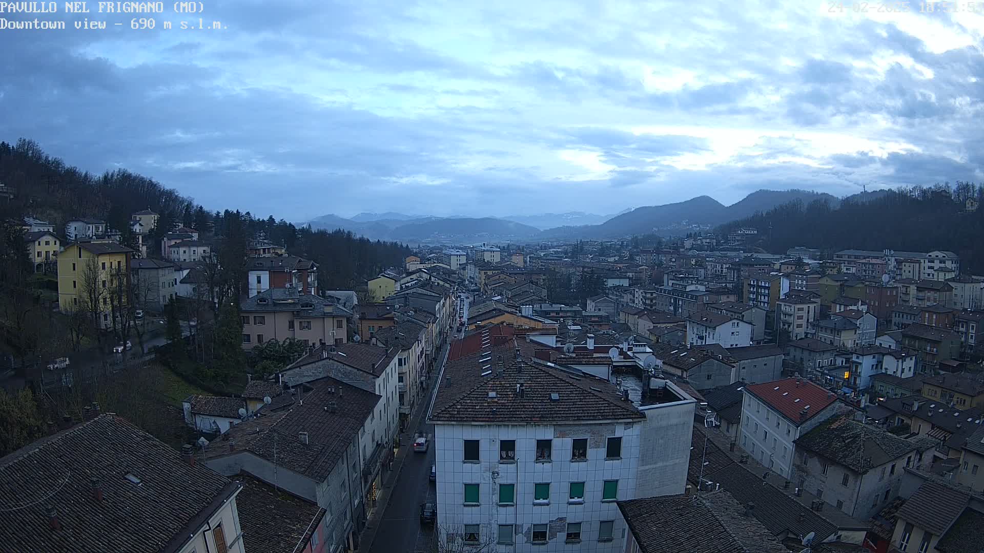 A sprawling town nestled in a valley under a cloudy sky, with mountains visible in the distance.