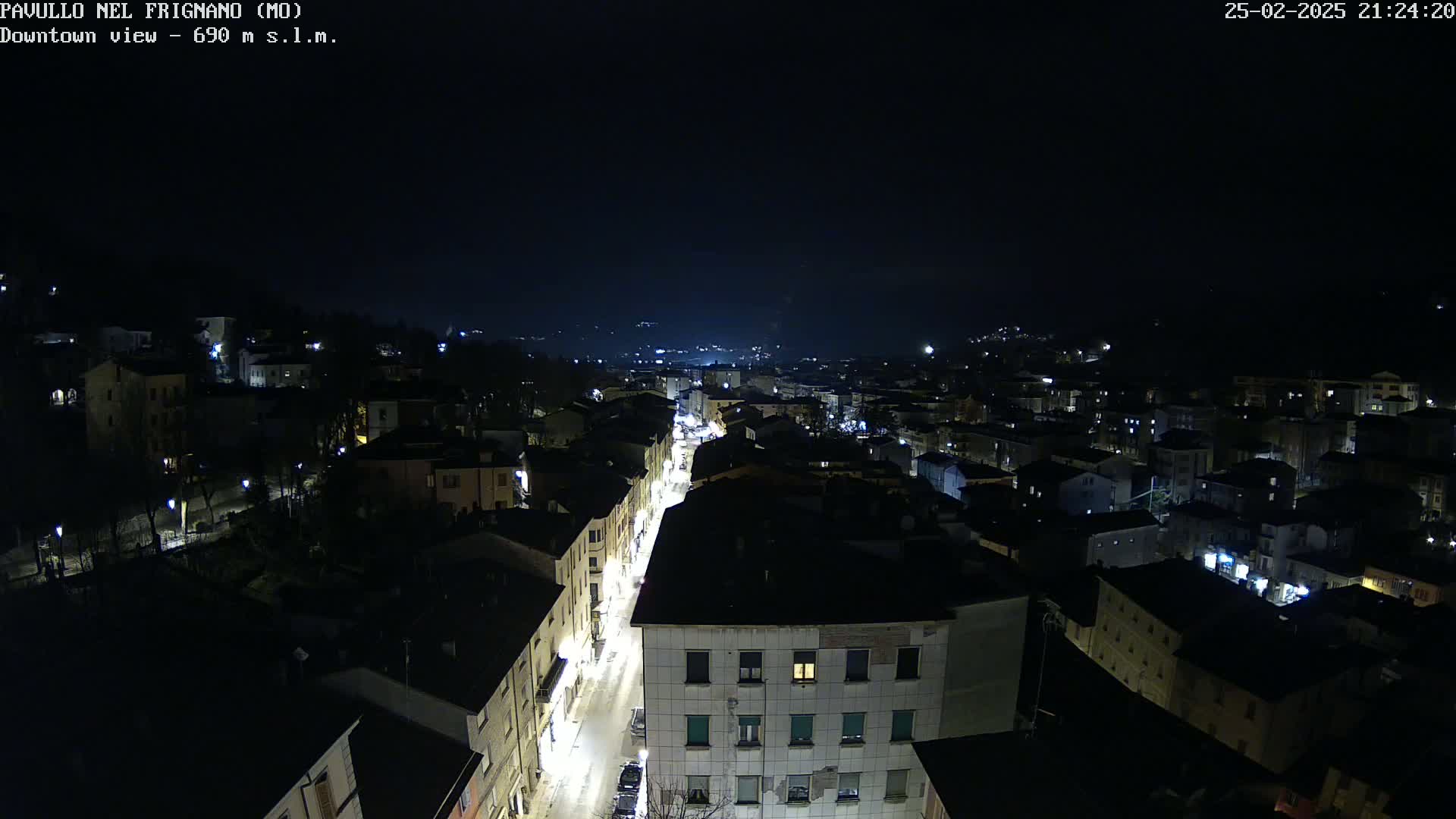 A nighttime aerial view of a town shows a street lined with illuminated buildings, extending into the dark distance under a clear, night sky.