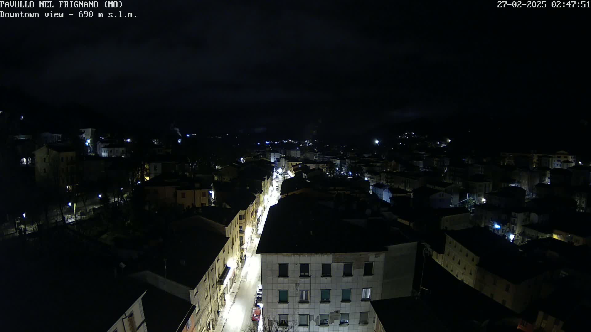 A nighttime, high-angle view of a town's streets and buildings illuminated by streetlights under a dark, cloudy sky.