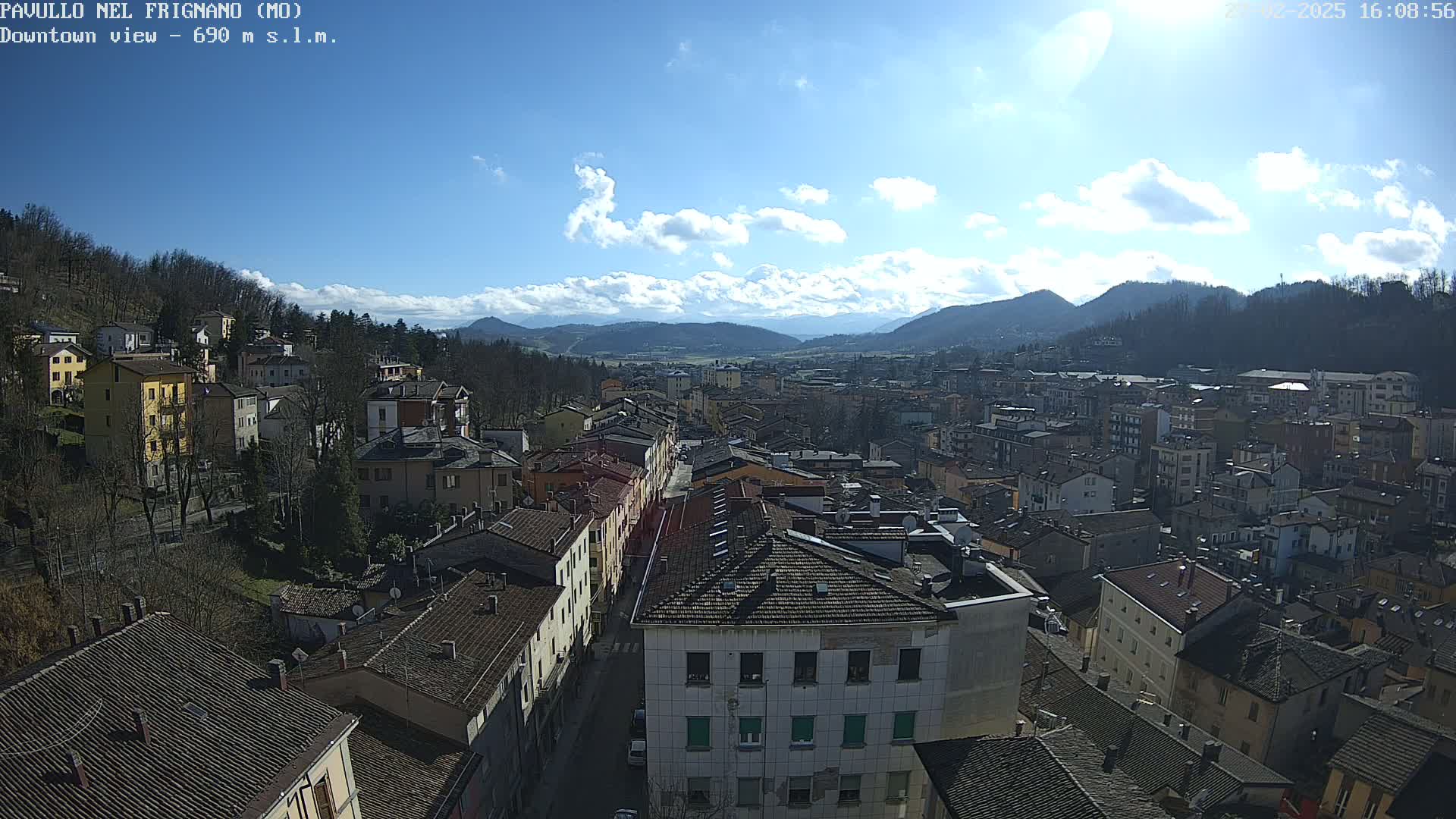 A sunny day reveals a sprawling town nestled in a valley, surrounded by mountains under a partly cloudy sky.