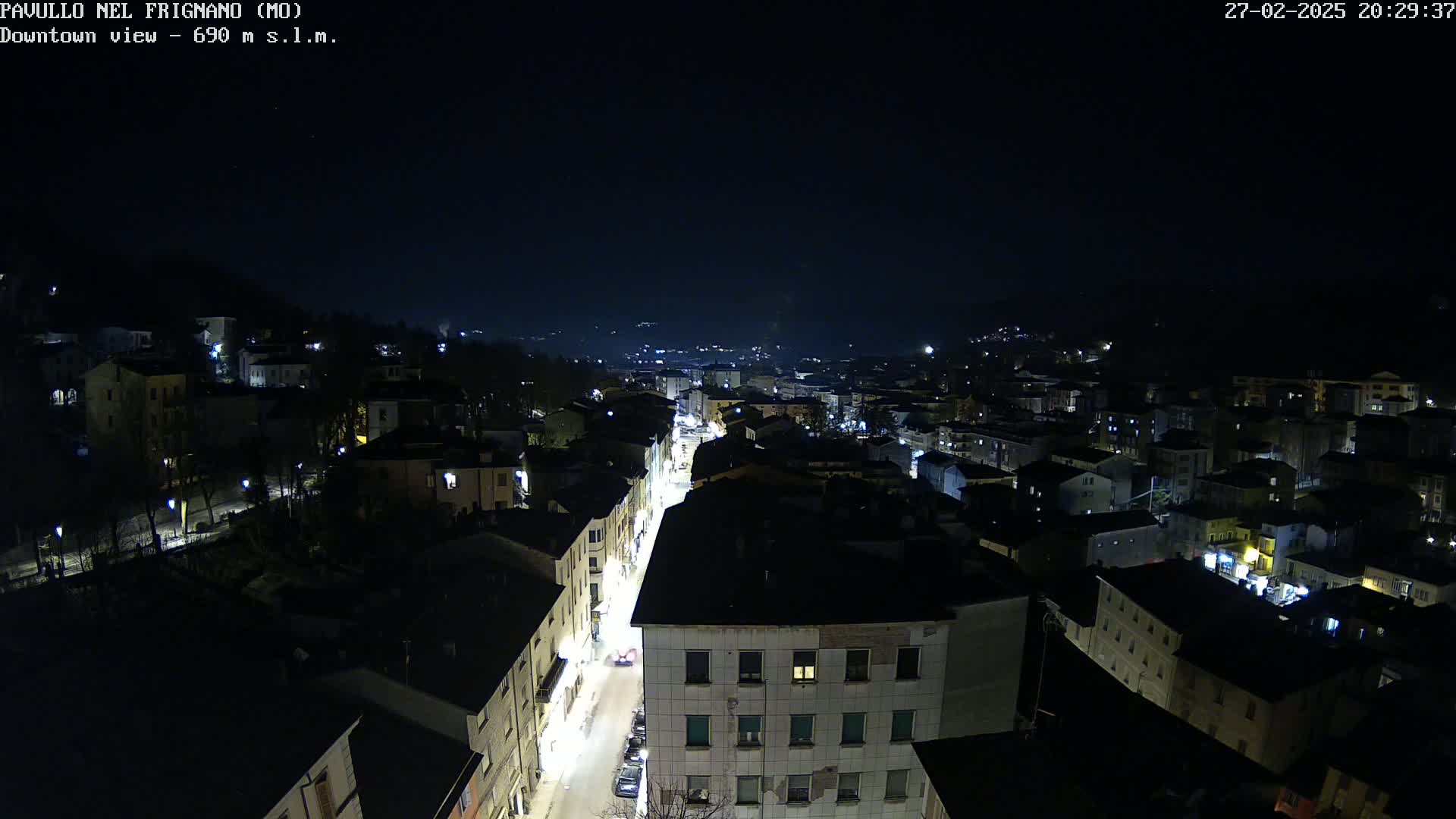 A nighttime aerial view of a town shows a long street with illuminated buildings and cars under a clear, dark sky.