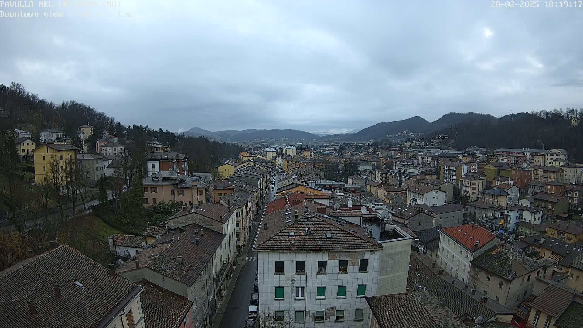 A high-angle, long shot of a town nestled in a valley on an overcast day, showing many buildings with red-tiled roofs.
