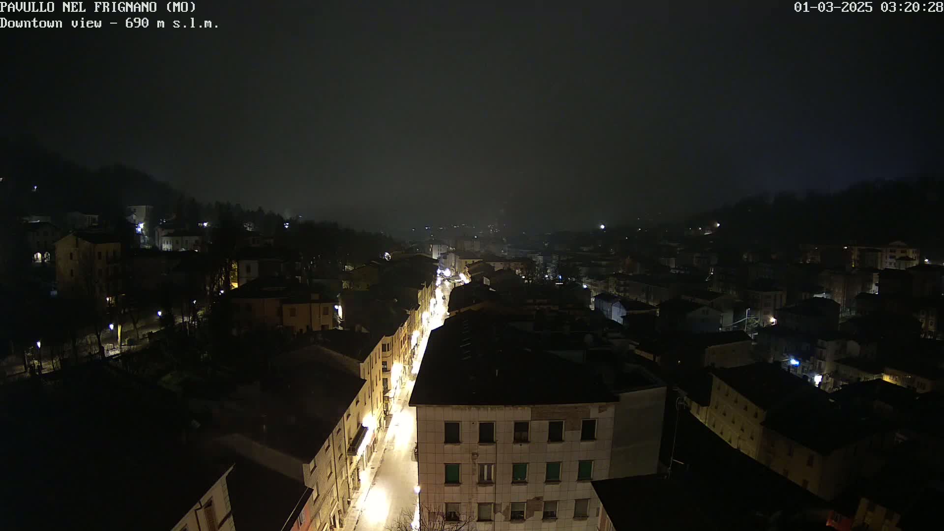 A nighttime, foggy view of a town shows a long, illuminated street winding through buildings, with hills visible in the distance.