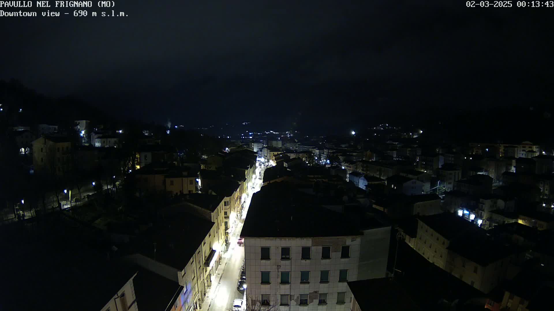 A nighttime, high-angle view shows a town with dimly lit streets and buildings under a dark, cloudy sky.