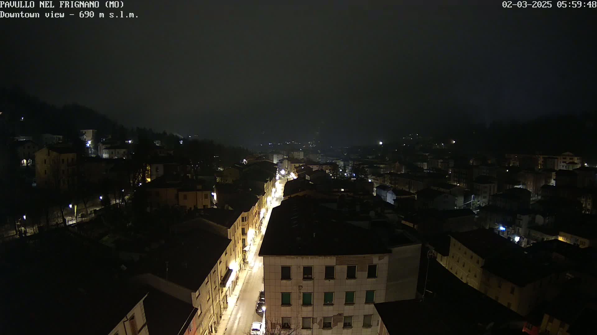 A nighttime, foggy view of a town shows a long, illuminated street cutting through a dense collection of dark buildings nestled against a hillside.