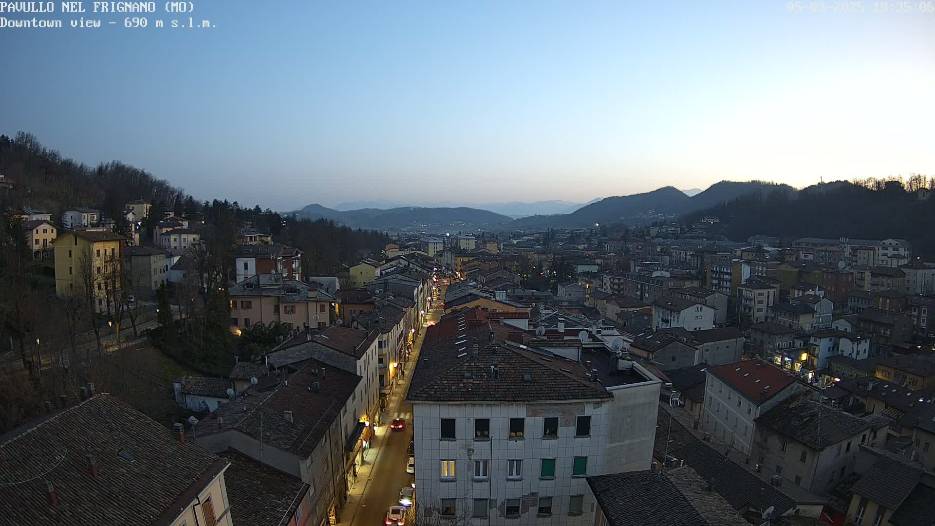 A sprawling town nestled in a valley at dusk, under a clear twilight sky, with a long street illuminated by streetlights.
