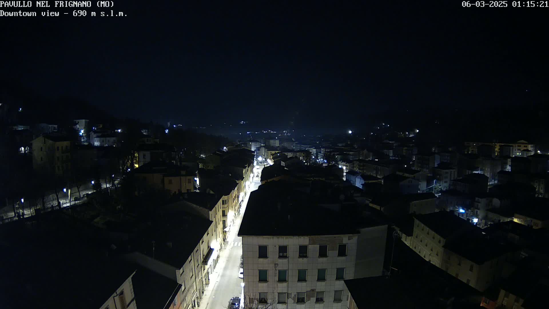 An aerial nighttime view shows a town with dimly lit buildings and a brightly lit street running through its center under a clear, dark sky.