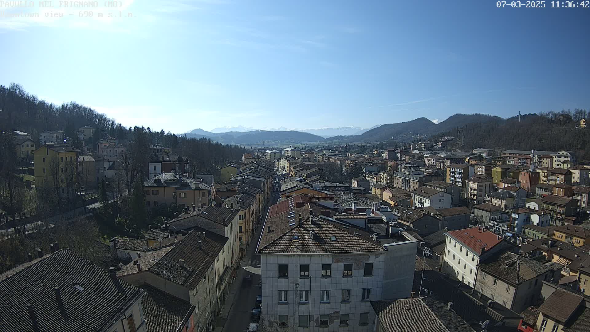 A sprawling town of closely packed buildings sits nestled in a valley, with snow-capped mountains visible in the distance under a clear blue sky.