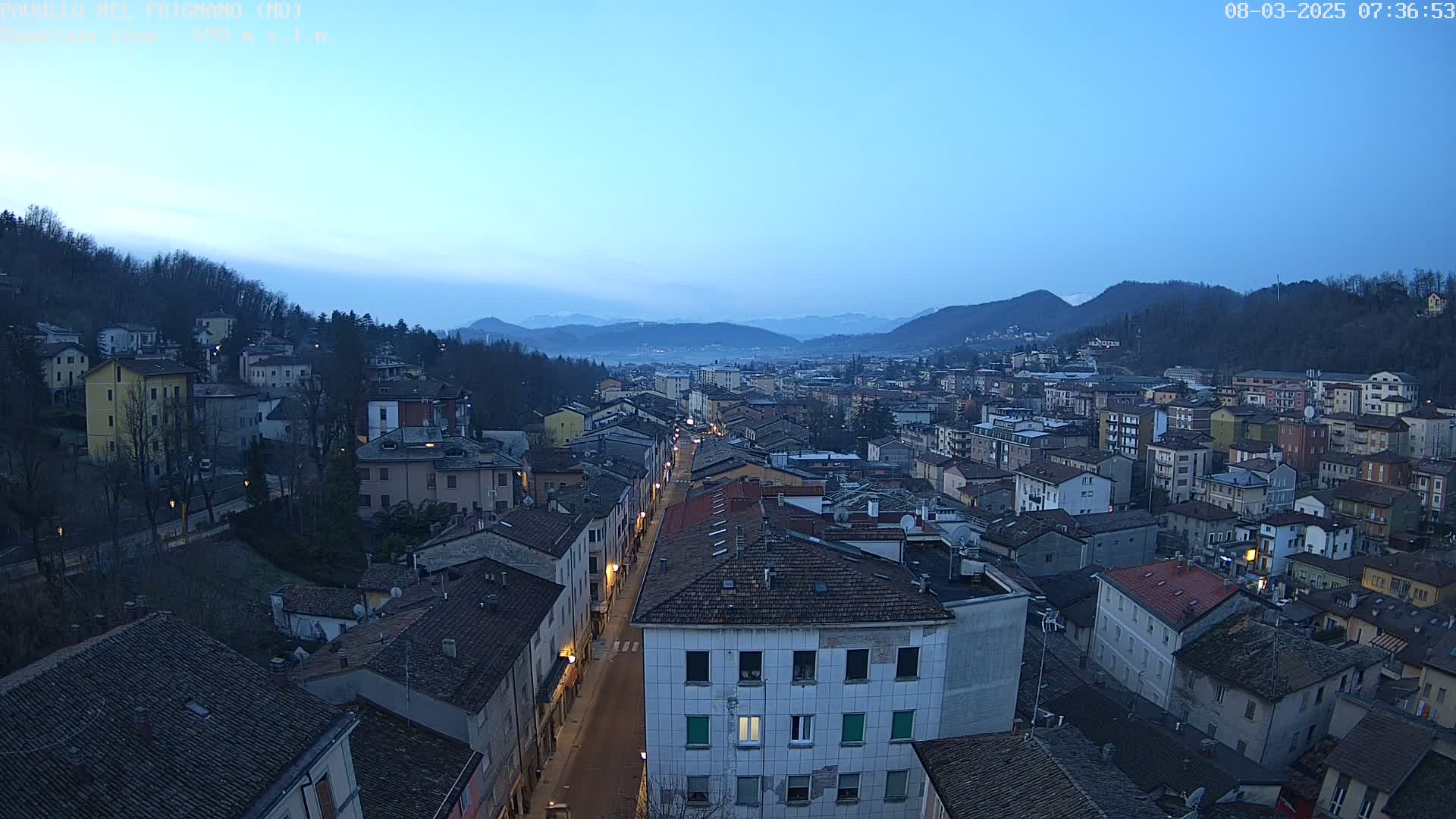 A long, straight street lined with buildings runs through a town nestled in a valley between hills under a twilight sky.