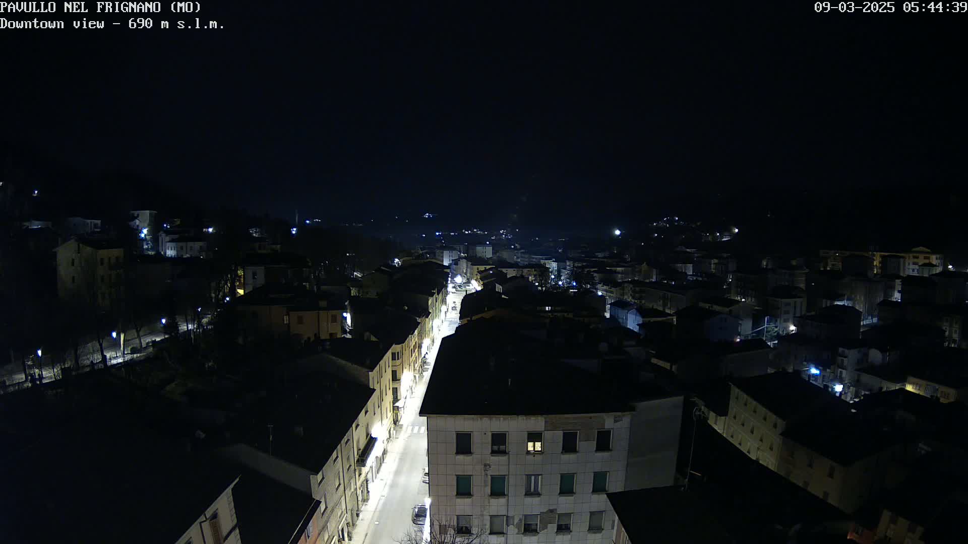 A nighttime, high-angle view shows a town's streetlights illuminating buildings and a road, surrounded by dark hills under a clear, black sky.