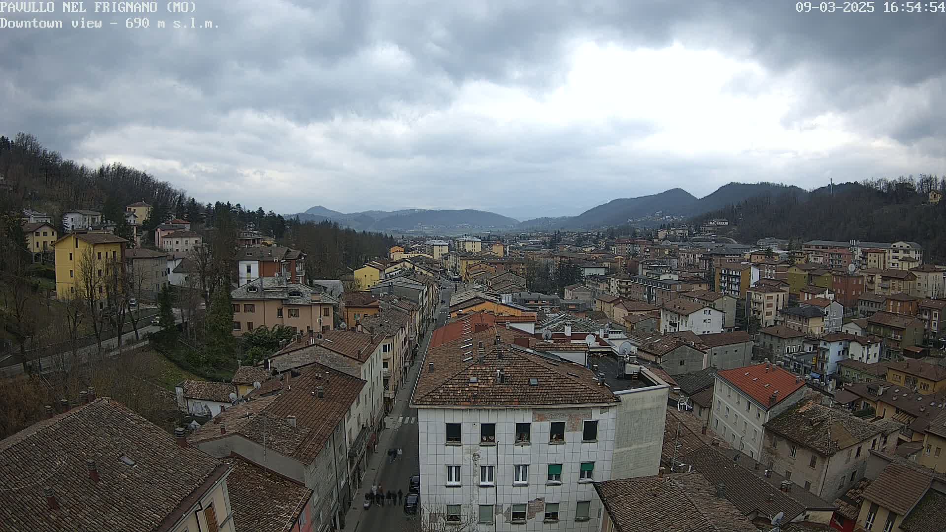 A view of a town nestled in a valley under a cloudy sky, with numerous buildings of varying heights and brown tiled roofs.