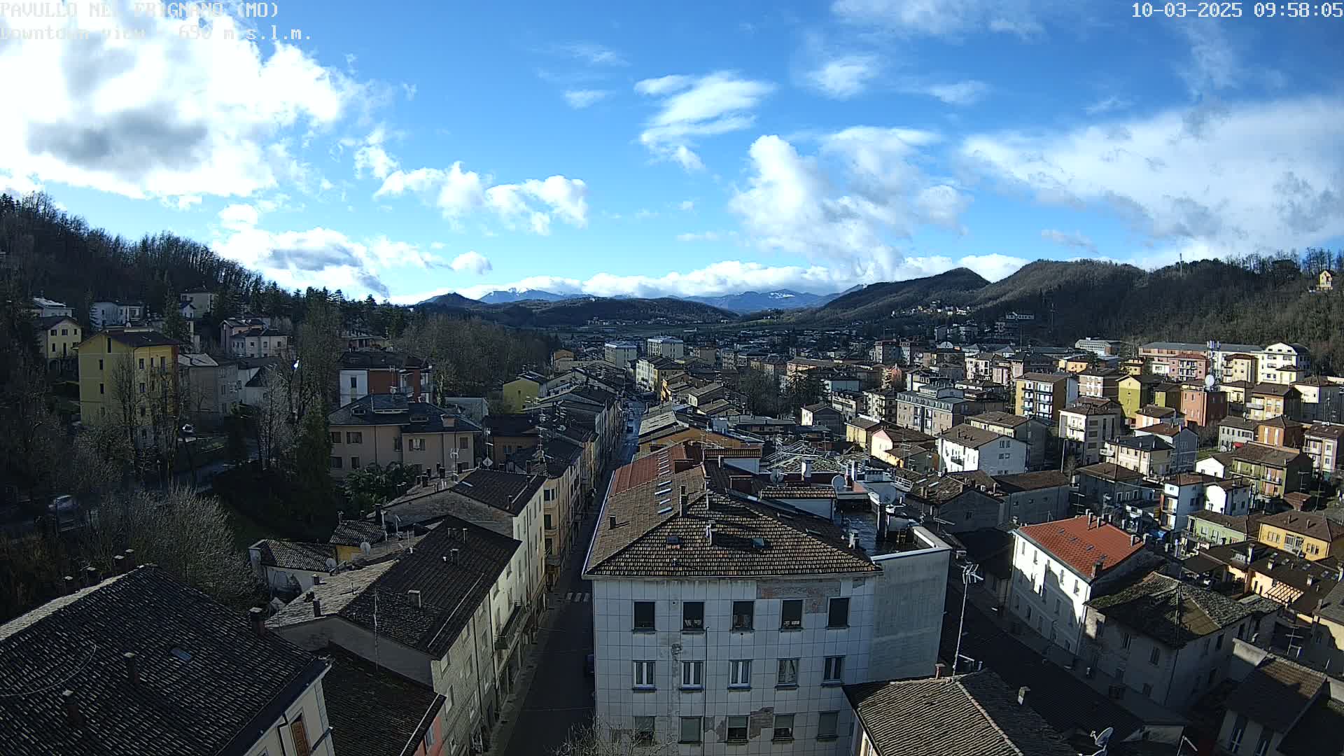 A partly cloudy day reveals a sprawling town nestled in a valley between tree-covered hills and distant mountains.