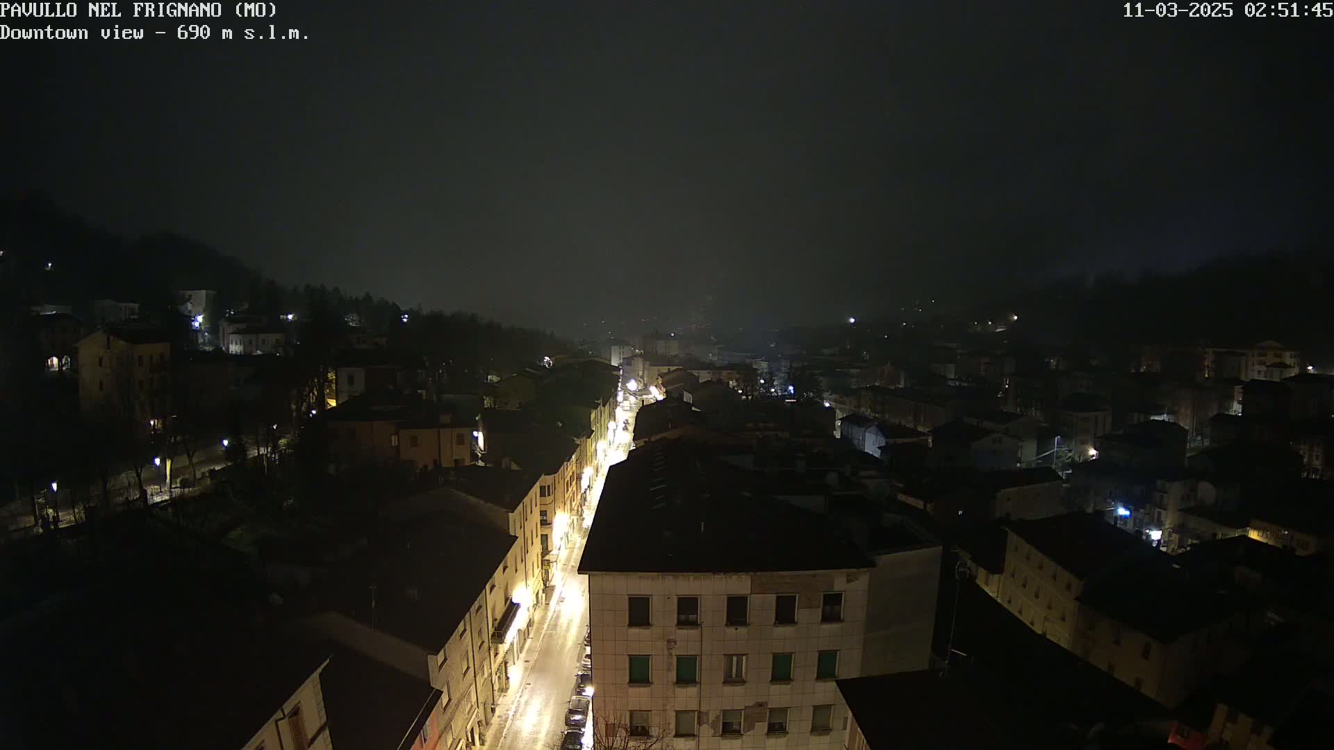 A nighttime, foggy view of a town's downtown area, with illuminated streets and buildings visible from an elevated perspective.