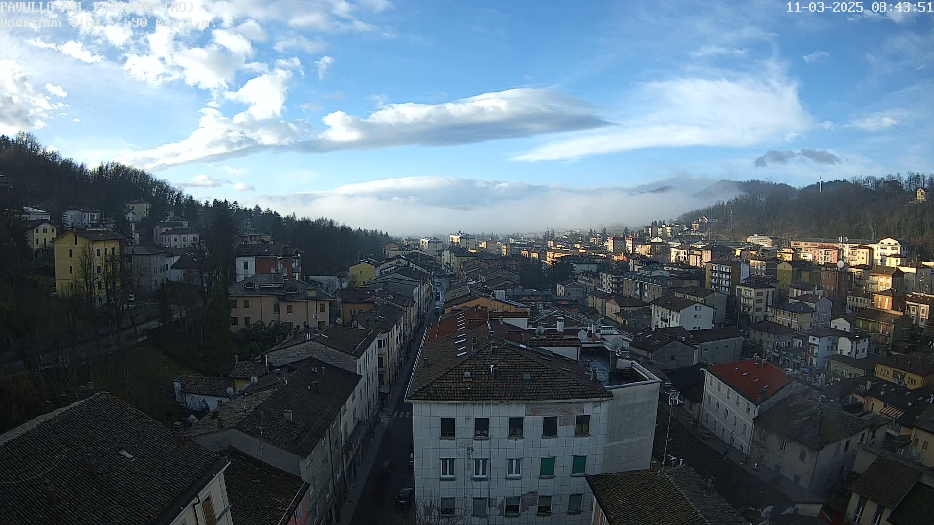 A mostly clear, blue sky with some clouds hangs over a dense collection of buildings in a valley, with fog settling in the distance on the hillsides.
