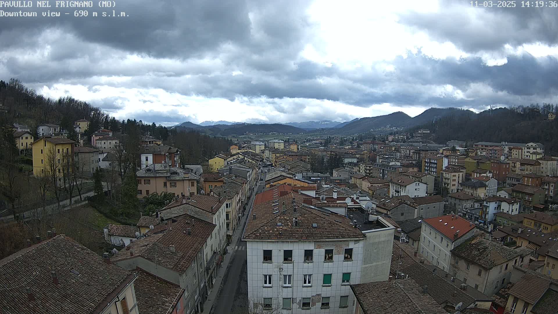 A high-angle view of a town nestled in a valley under a cloudy sky, showing a street running through the densely packed buildings and mountains in the distance.