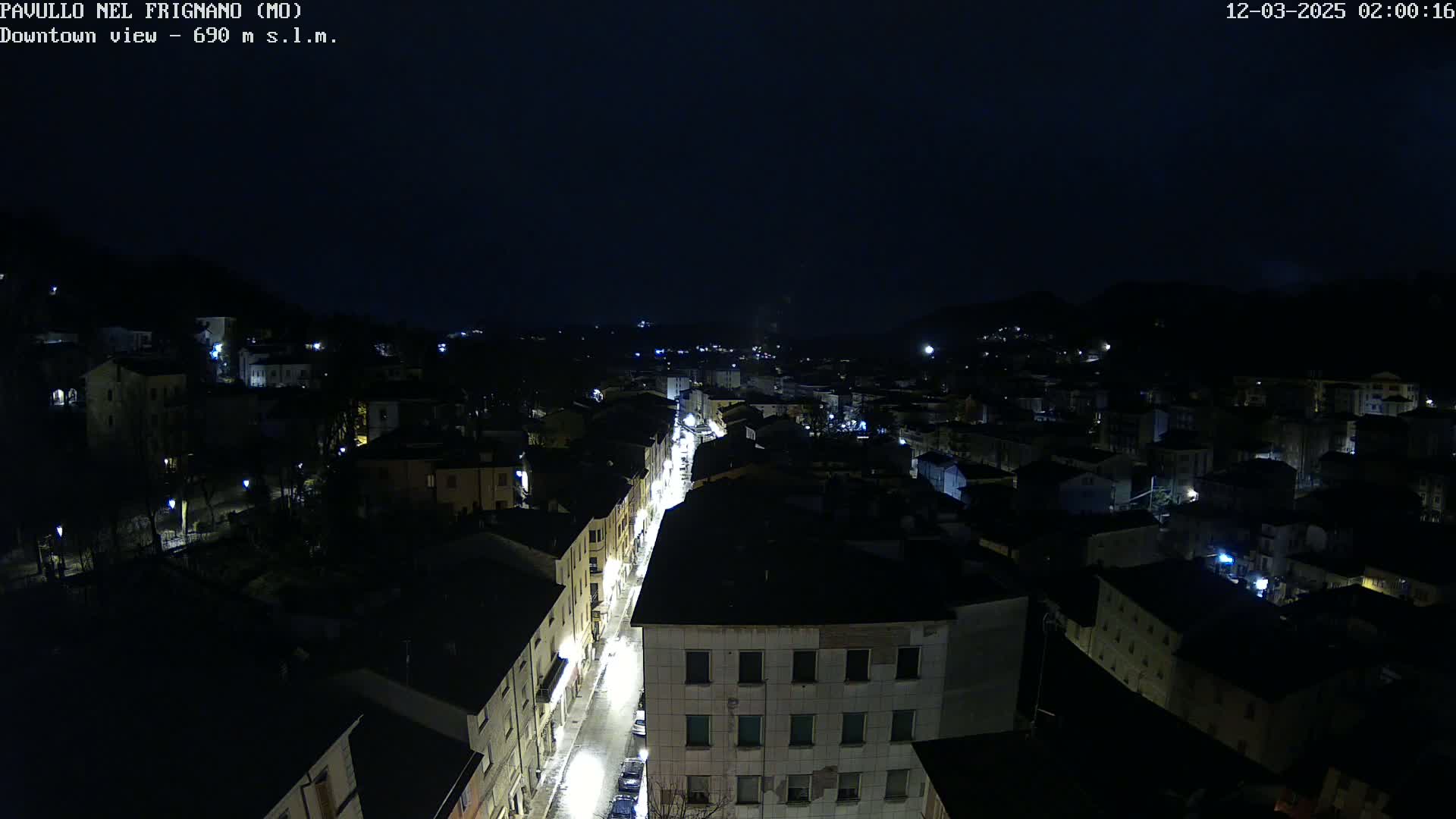A nighttime, high-angle view shows a town illuminated by streetlights and house lights, nestled in a dark, hilly landscape under a clear, dark sky.