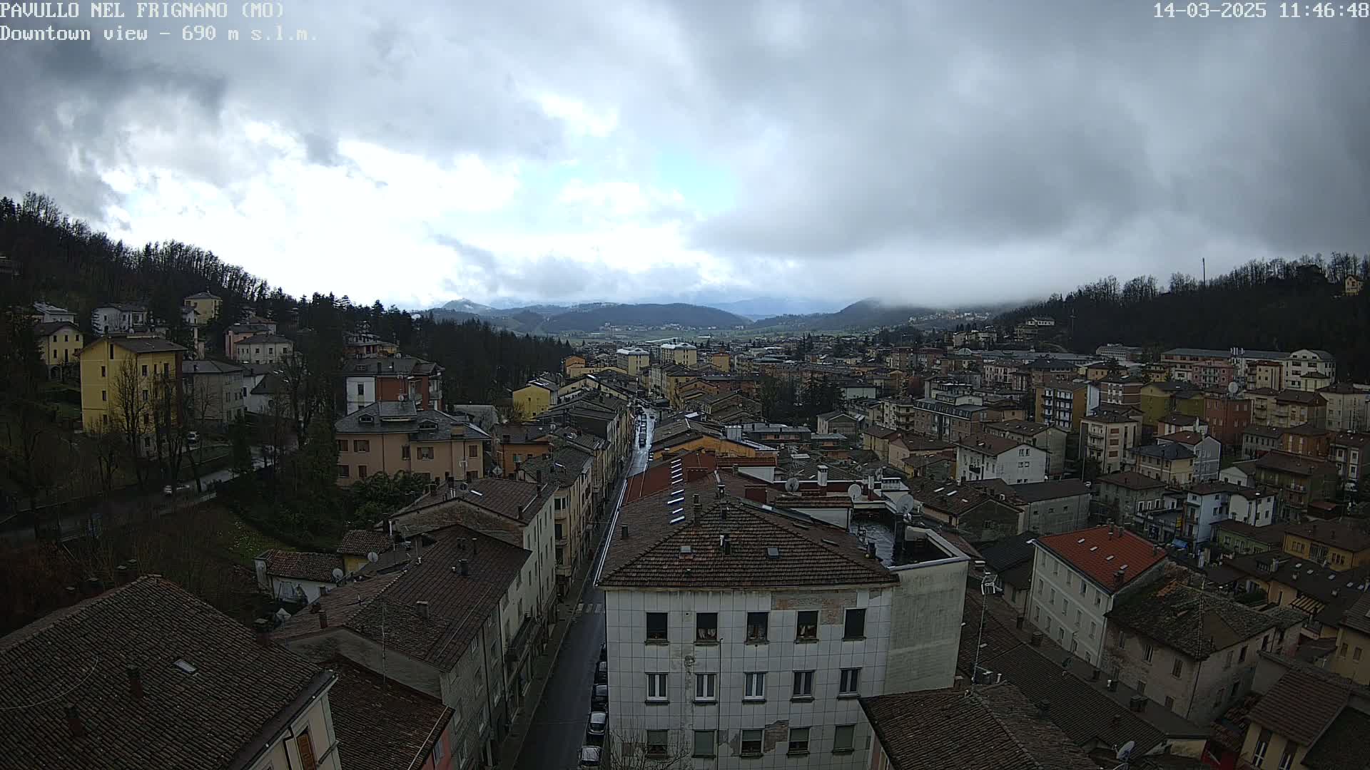 A high-angle view of a town on a cloudy day, showing a street running through the densely packed buildings, surrounded by hills and mountains in the distance.