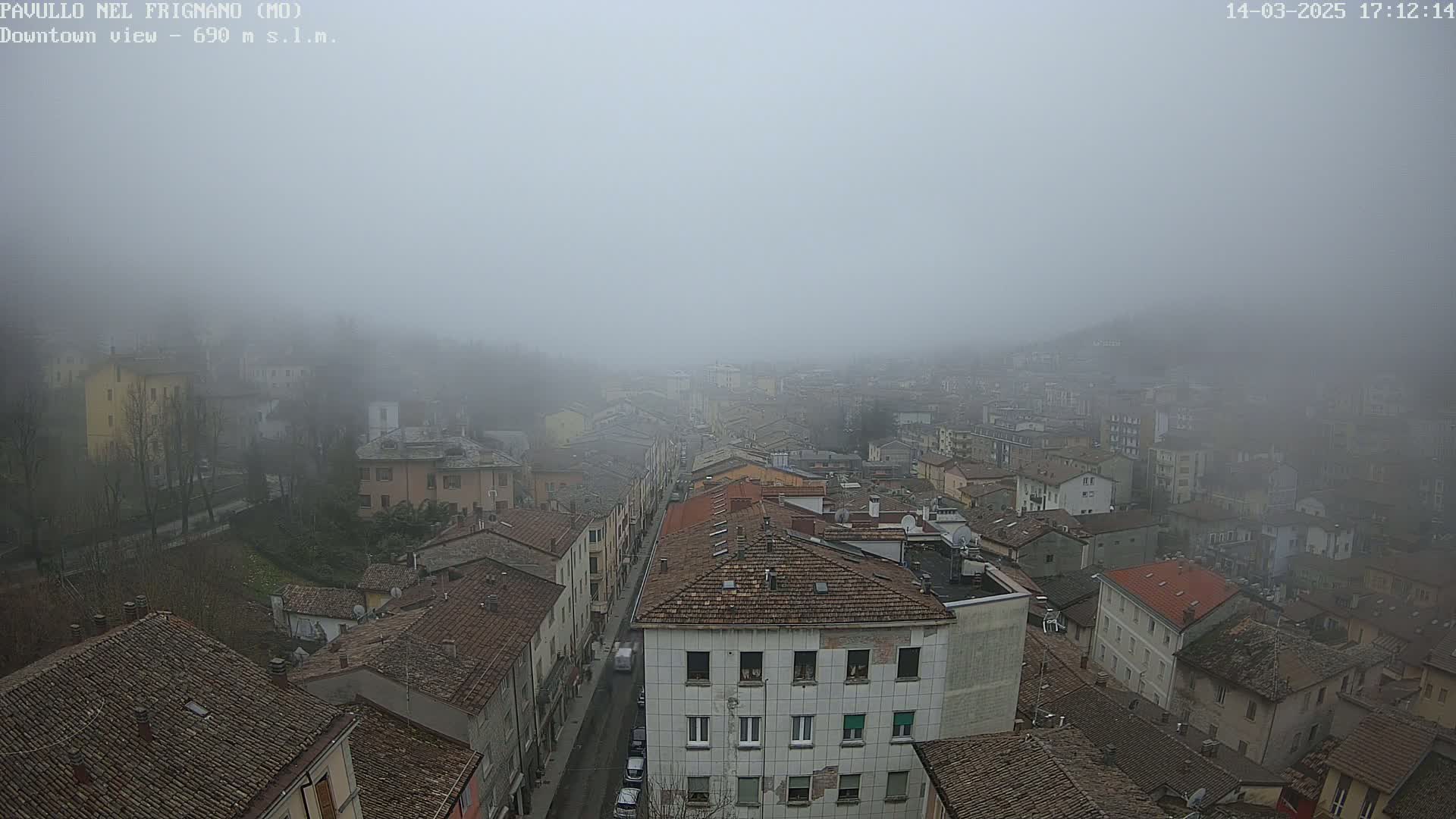 A foggy, high-angle view of a town's downtown area, showing a street lined with buildings that have red-tiled roofs.