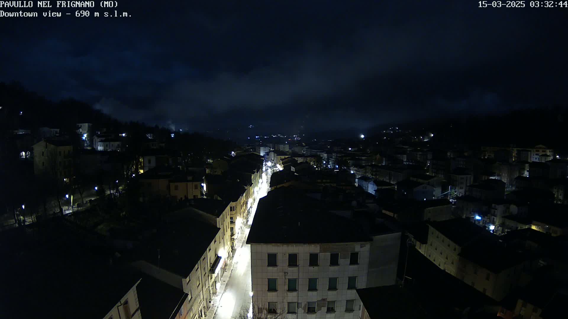 A nighttime, high-angle view of a town nestled in a valley under a cloudy sky, with a brightly lit street running through its center.