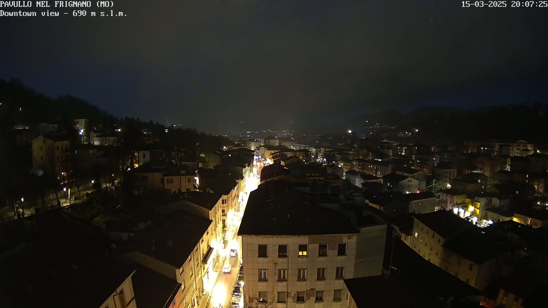 A nighttime aerial view of a town under a dark, possibly overcast sky, showing a brightly lit main street lined with buildings.