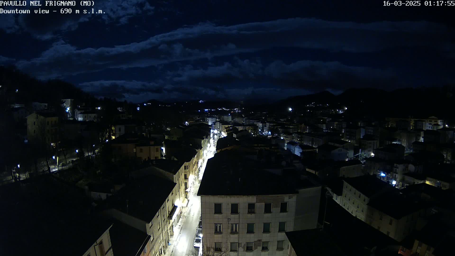 A nighttime aerial view of a town with a street lit by lamps under a partly cloudy sky.