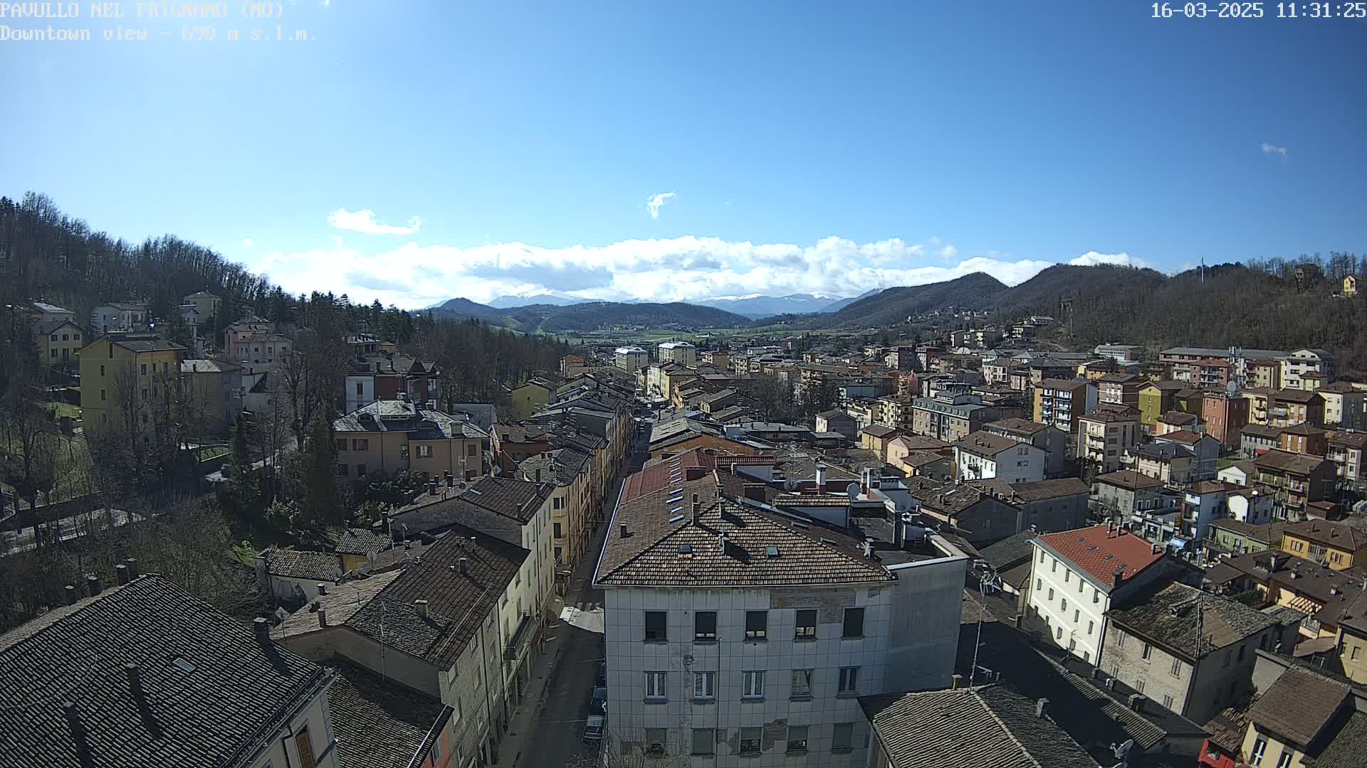A sunny day reveals a sprawling town nestled in a valley, surrounded by tree-covered hills and distant mountains under a partly cloudy sky.
