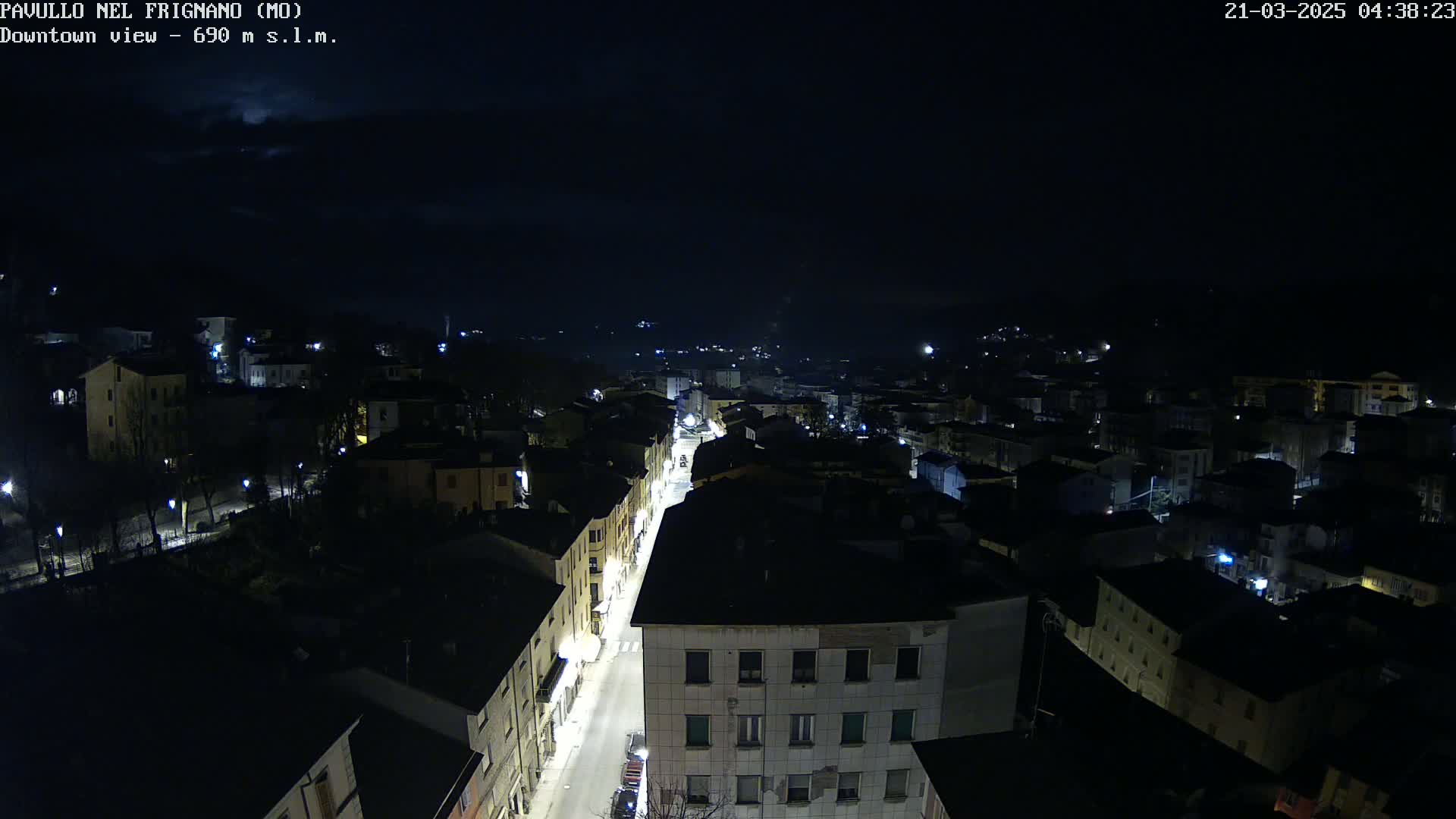 An aerial nighttime view of a town under a partly cloudy sky, showing a long street lit with streetlights extending from the foreground into the distance.