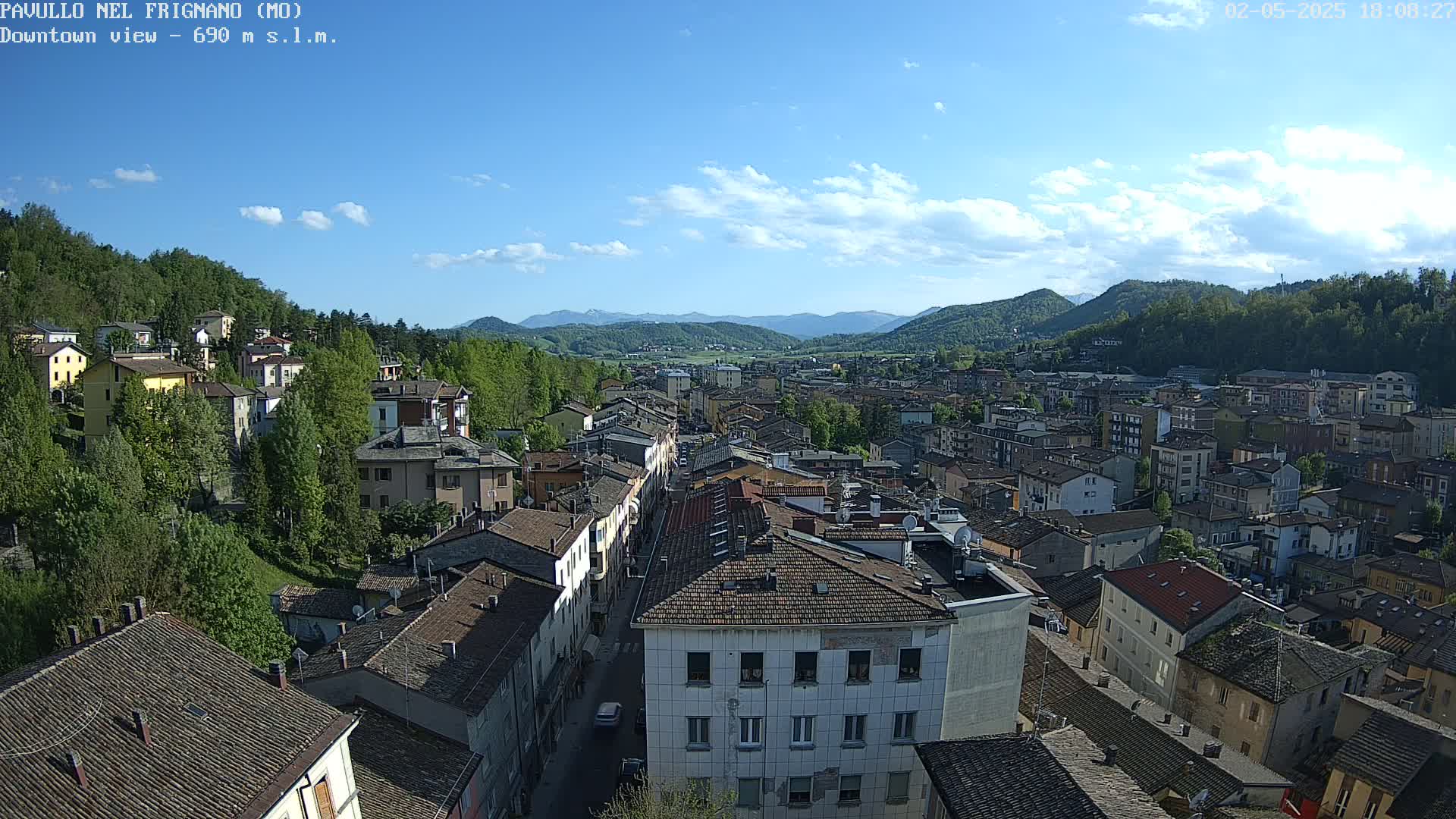 A sunny day reveals a sprawling town nestled in a valley, surrounded by lush green hills and mountains under a partly cloudy sky.