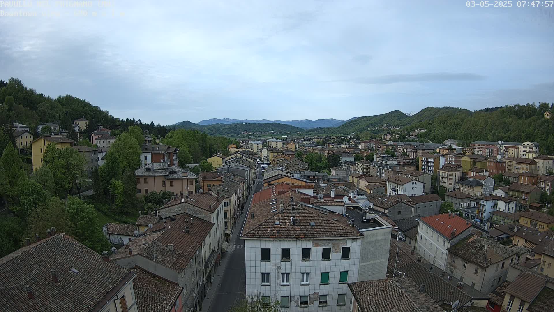 An elevated view of a town nestled in a valley under a mostly cloudy sky, surrounded by lush green hills and distant mountains.