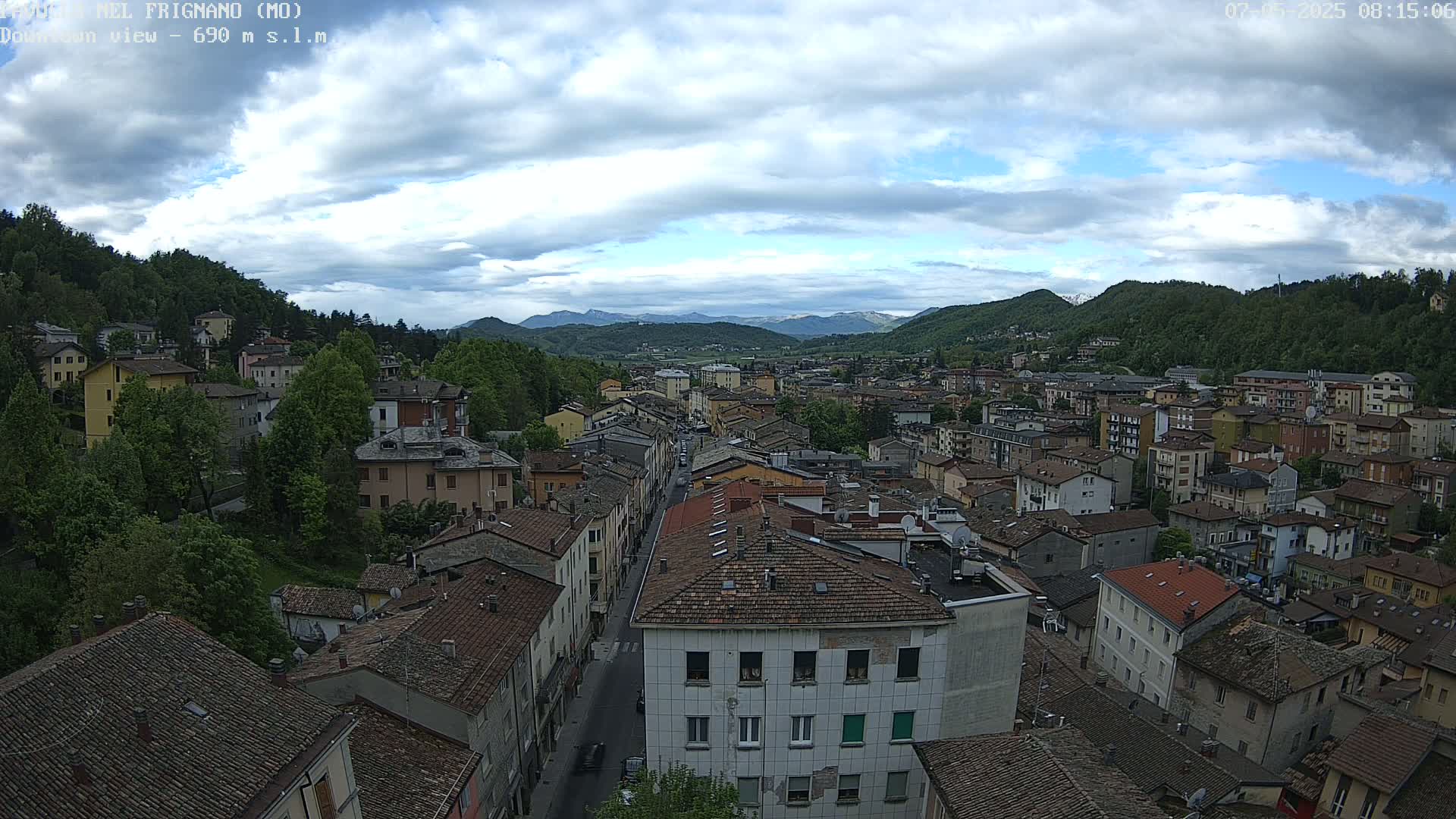 A partly cloudy day reveals a sprawling town nestled in a valley between verdant hills and distant mountains.