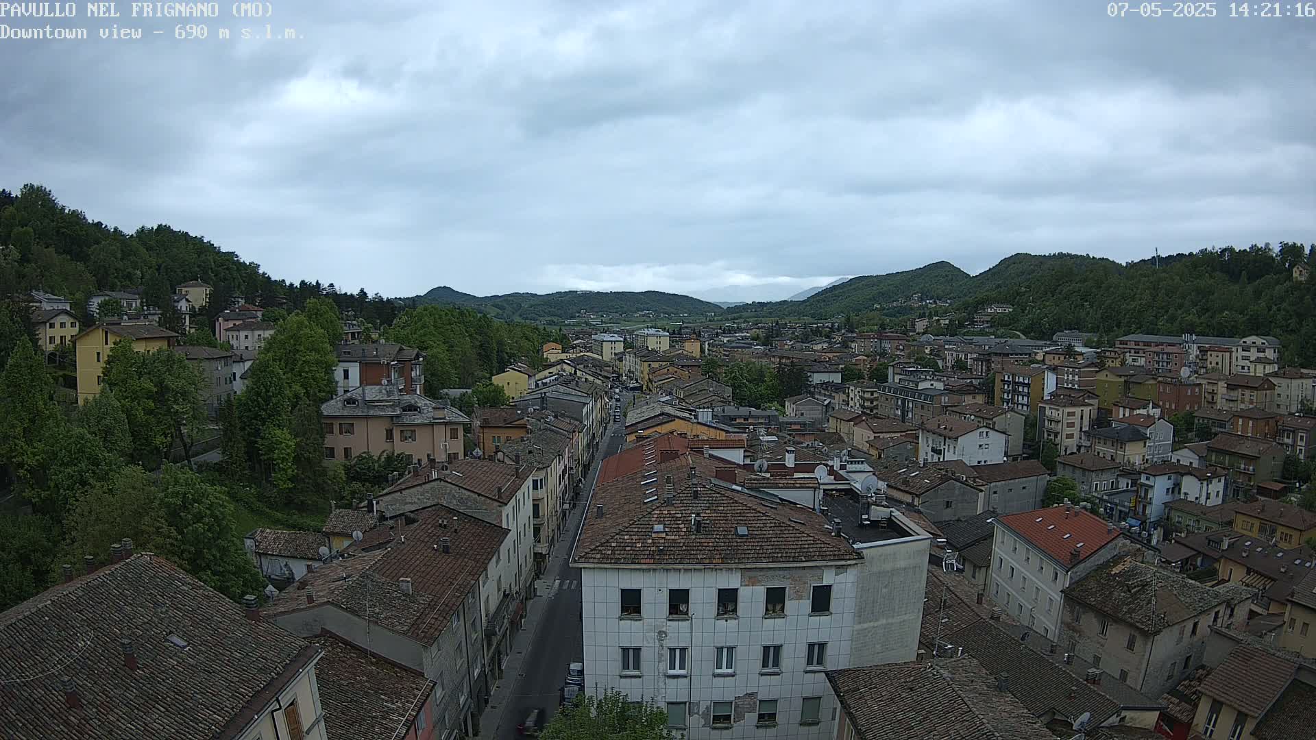 An elevated view of a town nestled in a valley under a cloudy sky, showing numerous buildings with red-tiled roofs and lush green hills in the background.