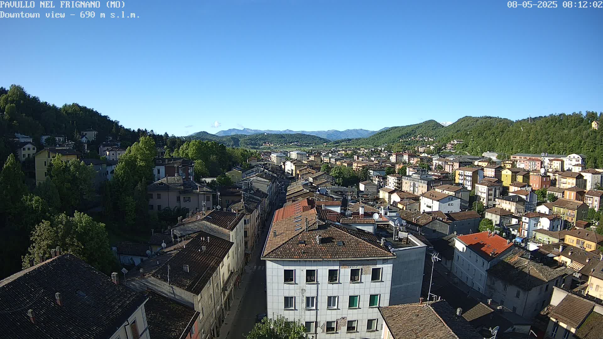 A high-angle view on a clear day shows a town nestled in a valley between green hills and mountains.