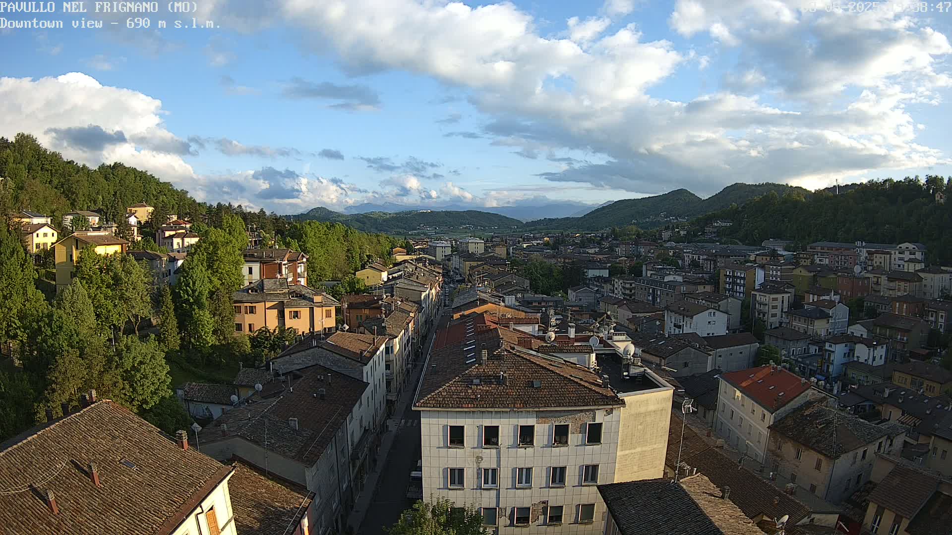 A partly cloudy afternoon view of a town nestled in a valley between hills, with many brown-roofed buildings stretching down a long street.