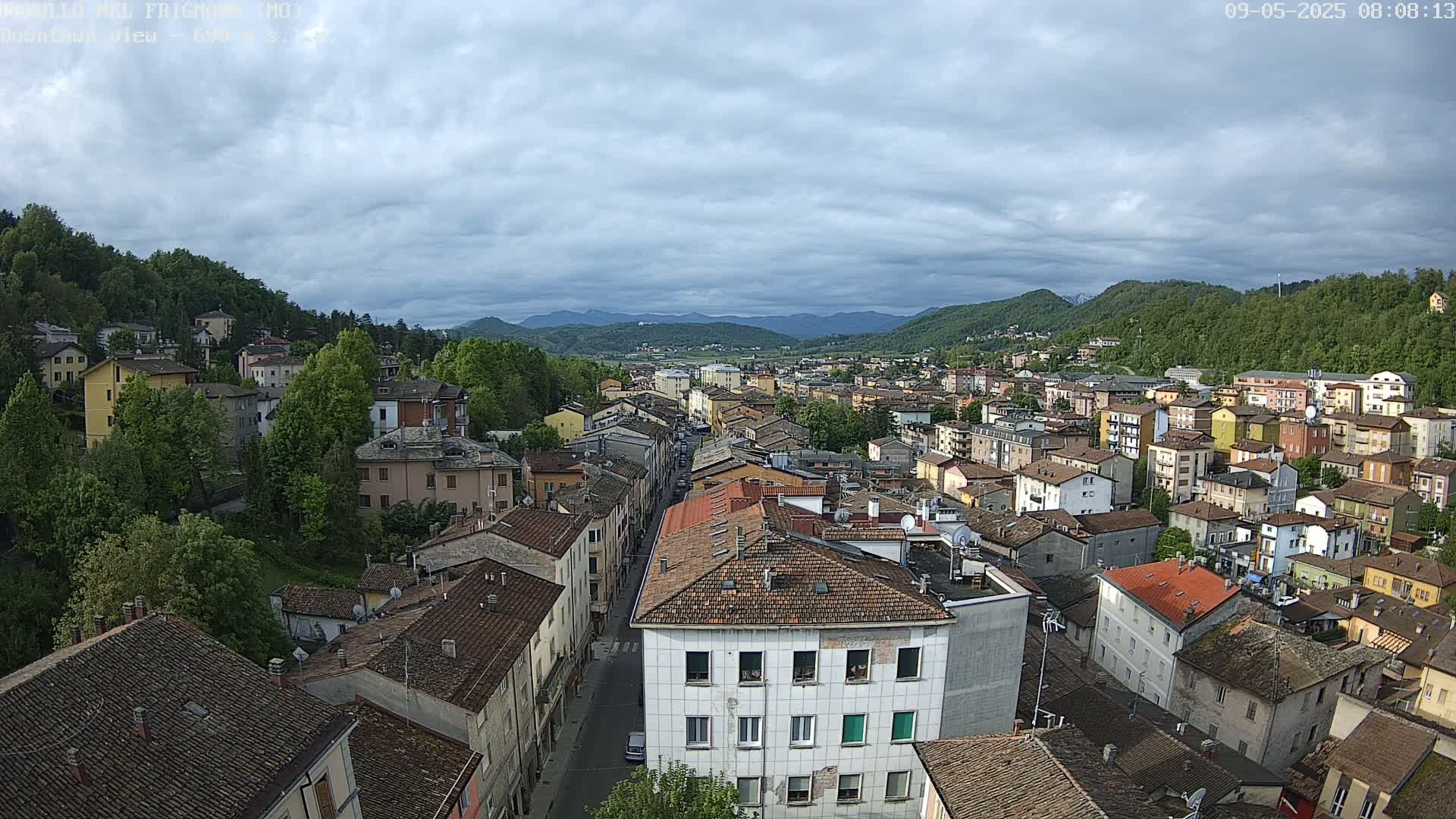 An elevated view of a town nestled in a valley, under a mostly cloudy sky, shows many buildings with red-tiled roofs.