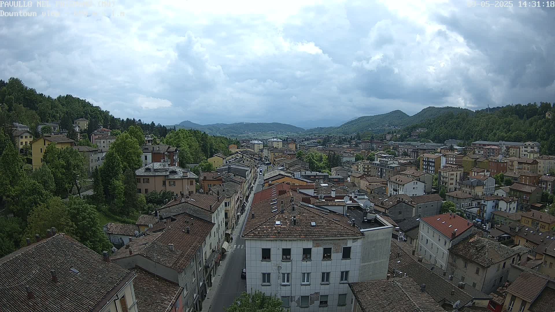 An elevated view of a town nestled in a valley under a partly cloudy sky shows numerous buildings with reddish-brown roofs, a long street running through the center, and green hills in the background.