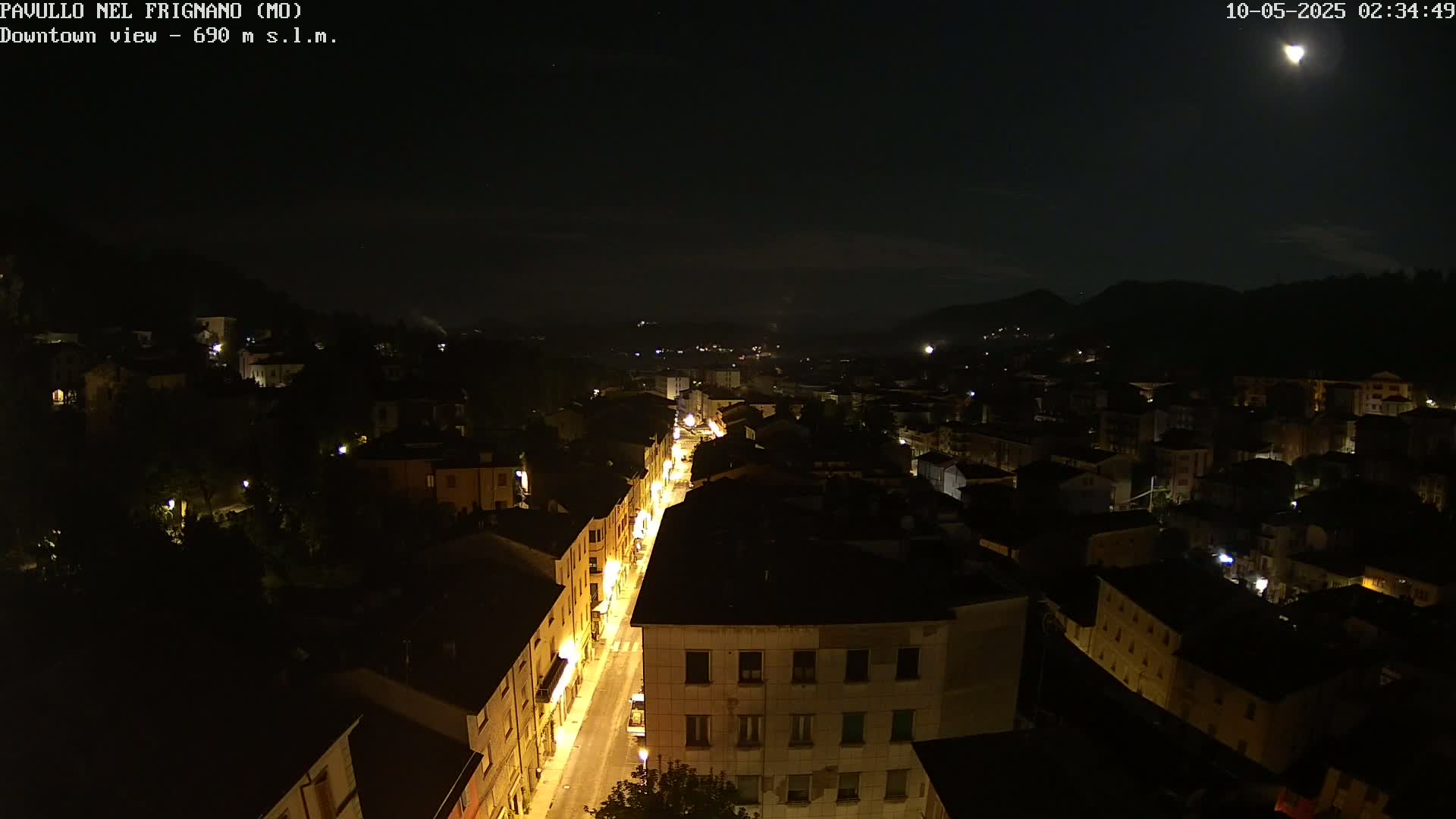A nighttime, high-angle view of a town illuminated by streetlights, with a bright moon visible in the sky and dark hills in the background.
