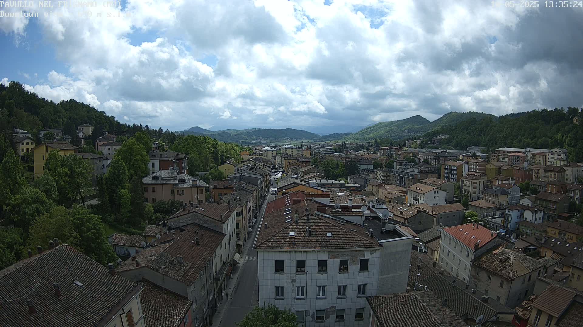 A partly cloudy day overlooks a town nestled in a valley between green hills.