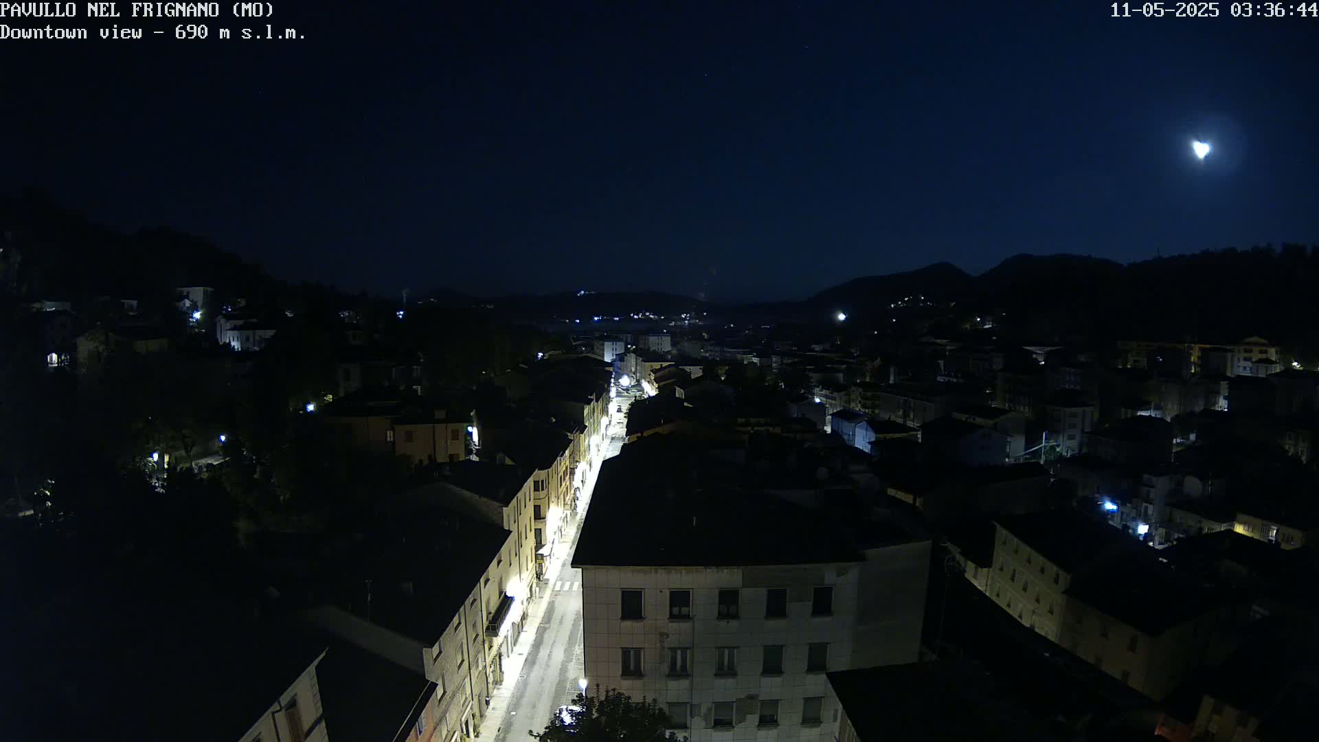 A nighttime, high-angle view of a town nestled between hills under a dark sky illuminated by a bright moon.
