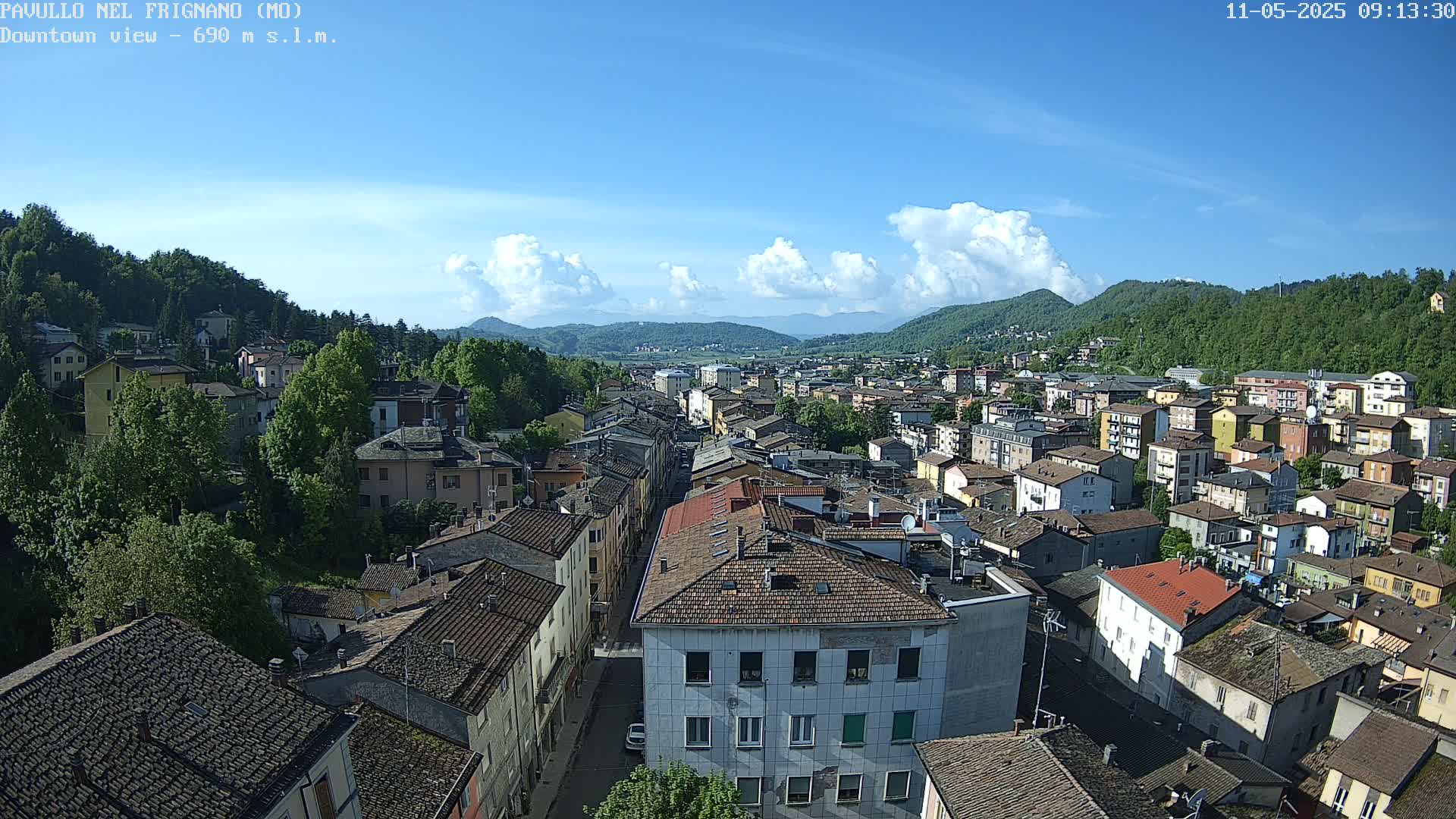 An aerial view of a town nestled in a valley, under a mostly clear blue sky with some cumulus clouds.