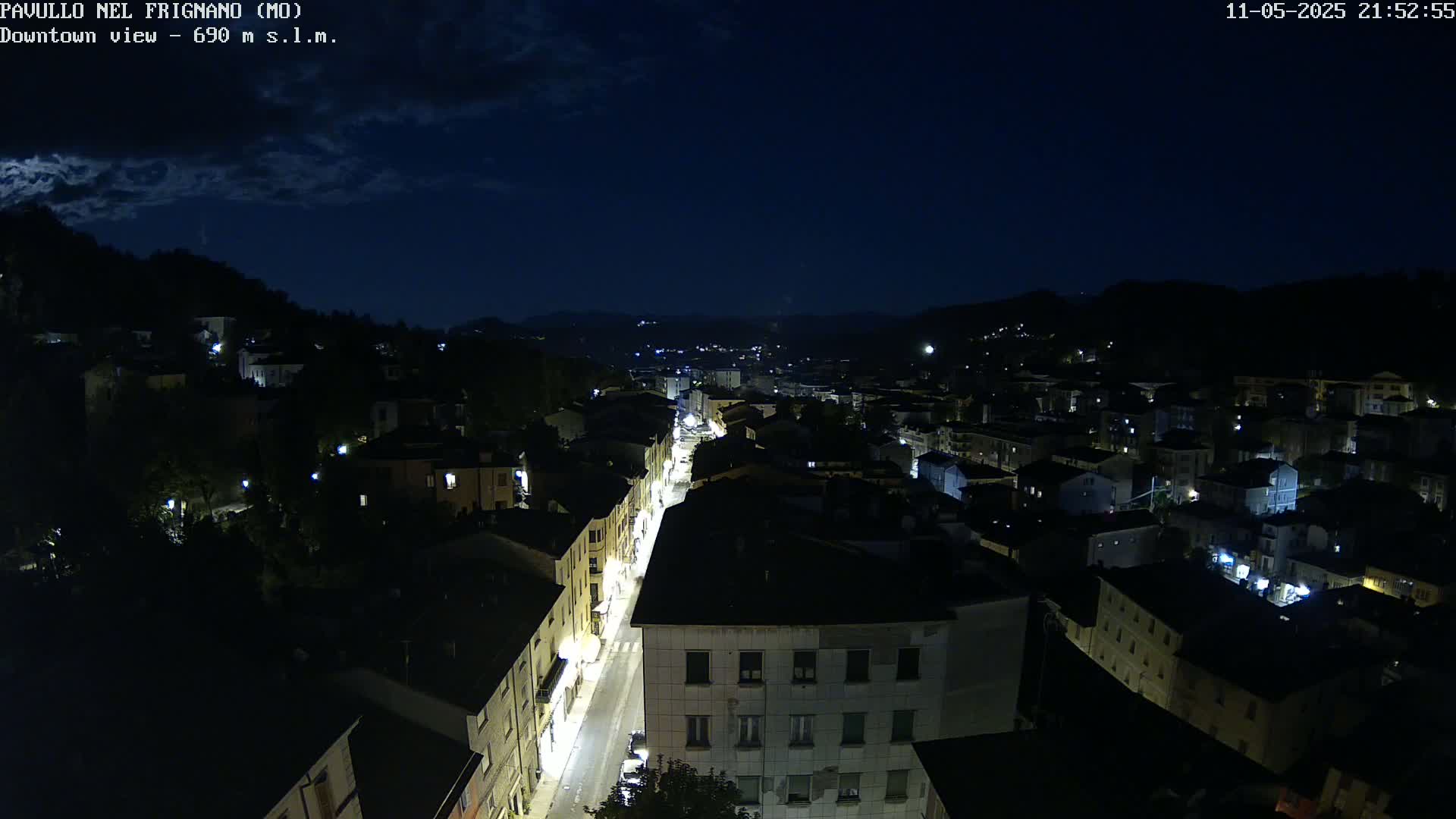 A nighttime, high-angle view of a town's illuminated streets and buildings under a partly cloudy sky.
