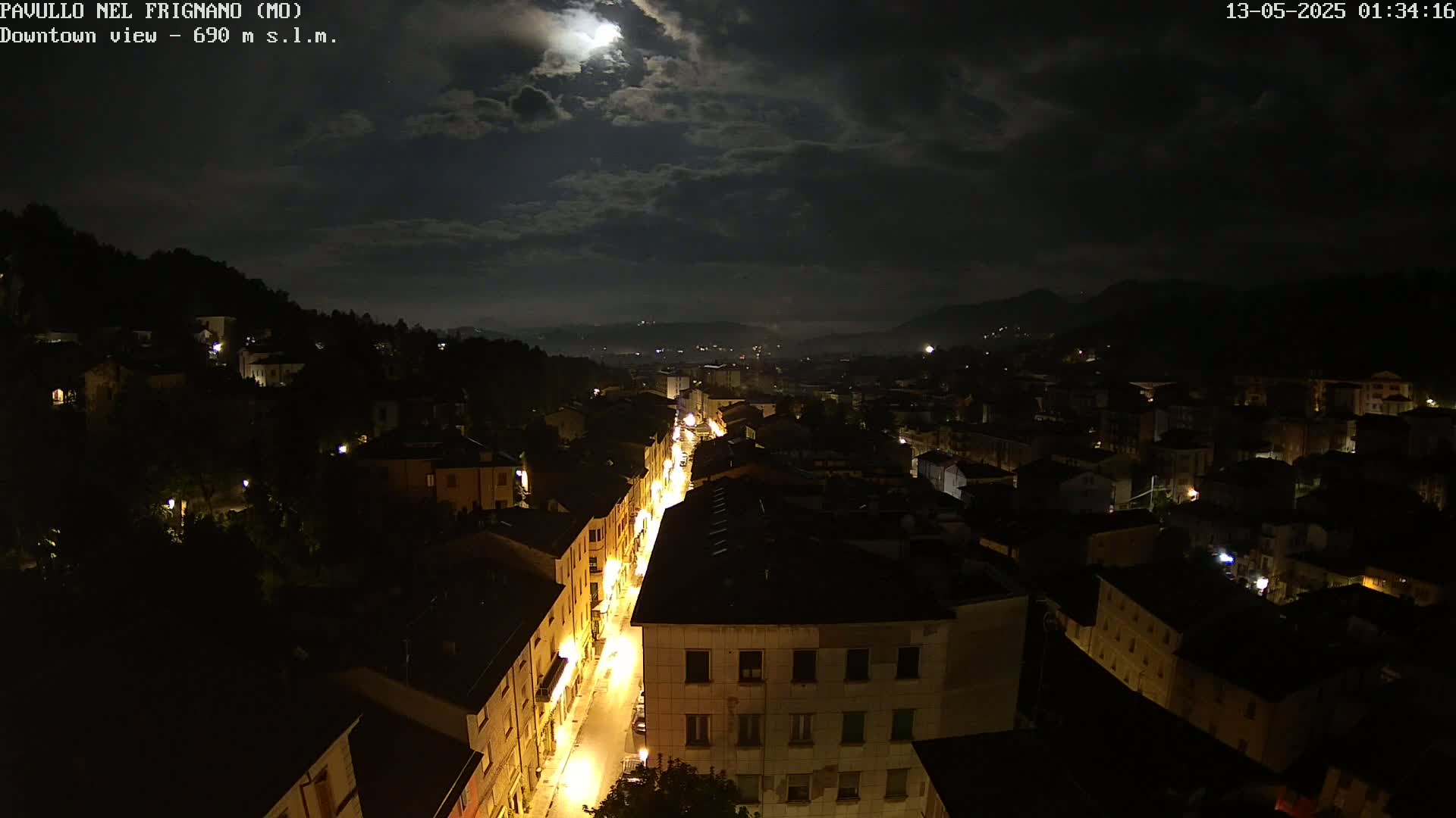 A nighttime, moonlit view of a town nestled in hills, with a brightly lit street winding through its center.