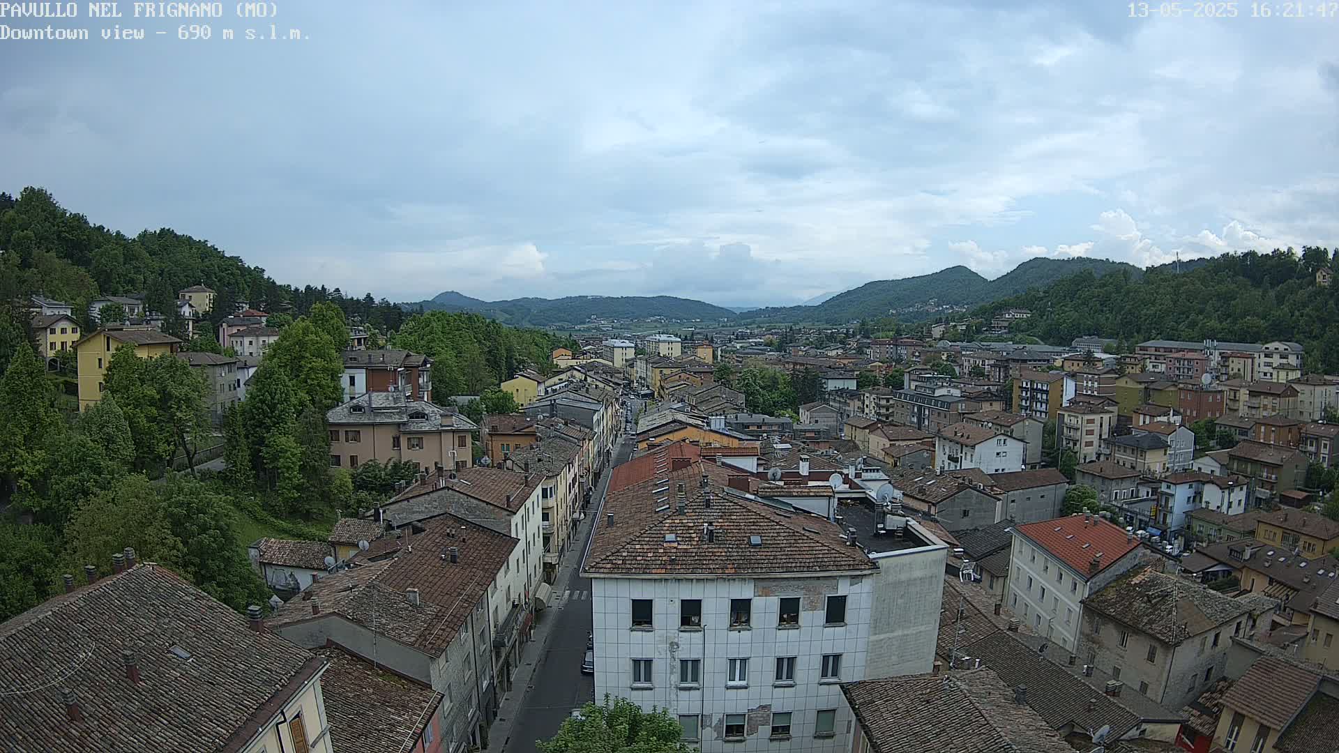 A mostly cloudy sky overlooks a dense collection of buildings nestled in a valley surrounded by lush green hills.