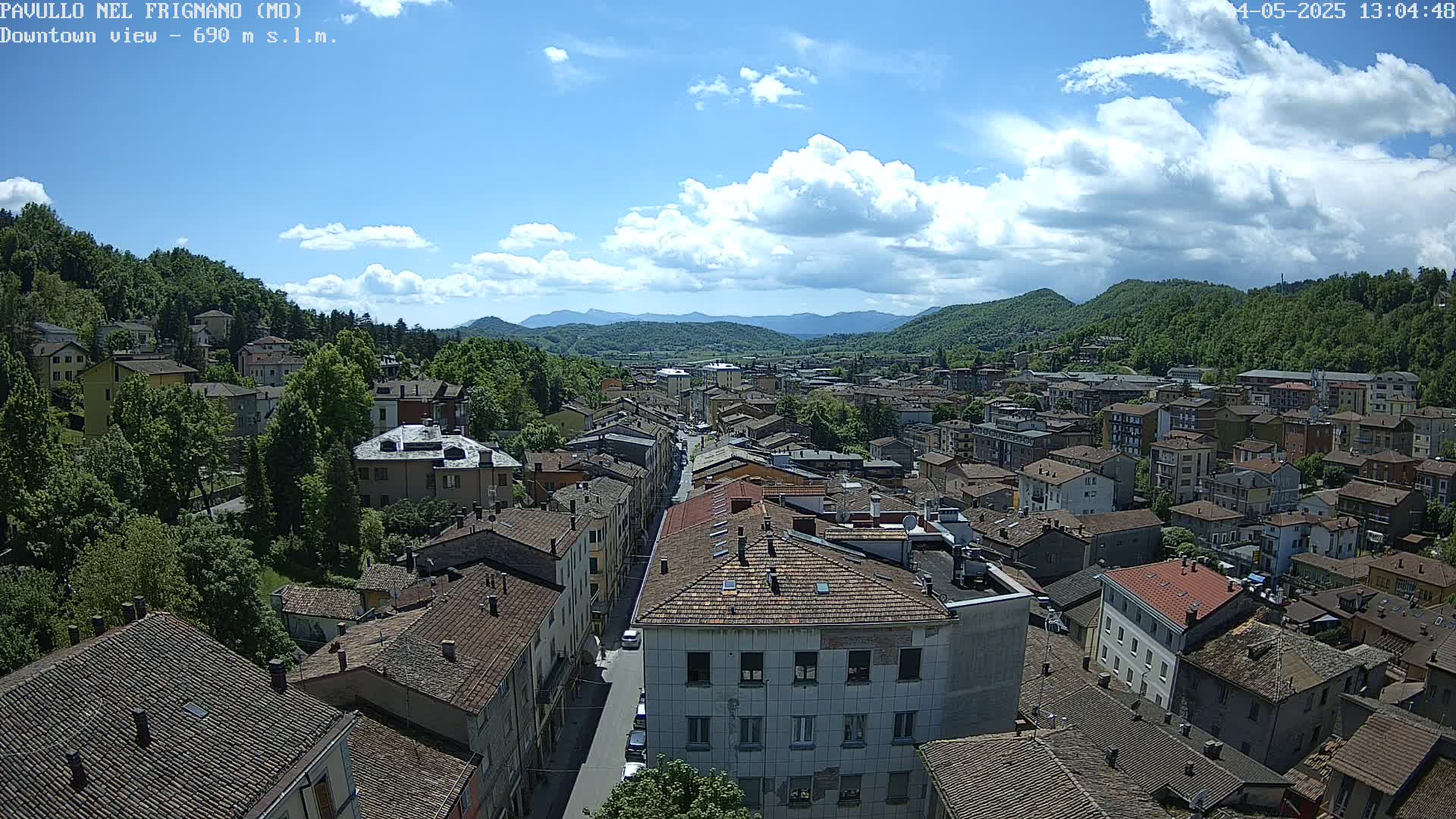 A sunny day reveals a sprawling town nestled in a valley, surrounded by lush green hills and mountains under a partly cloudy sky.
