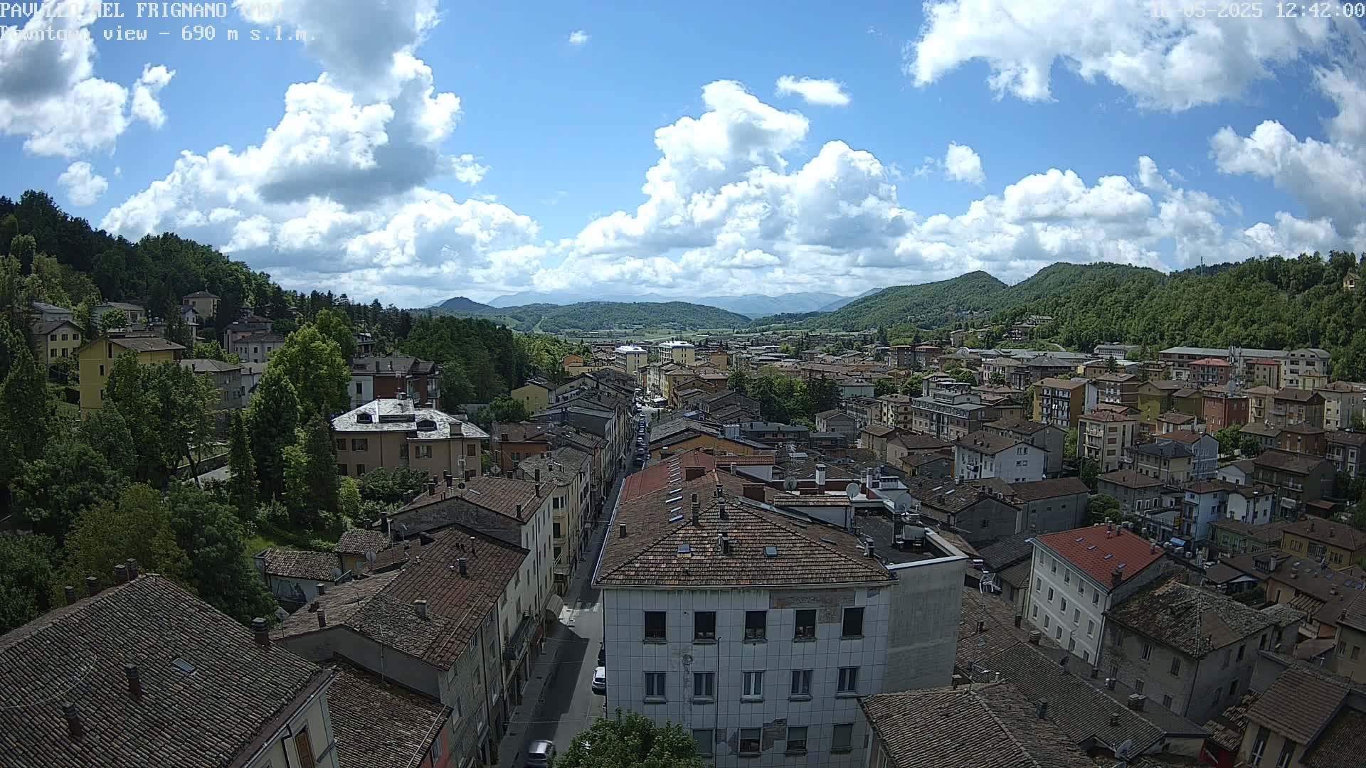 A partly sunny day reveals a panoramic view of a town nestled in a valley, surrounded by lush green hills and mountains under a partly cloudy sky.