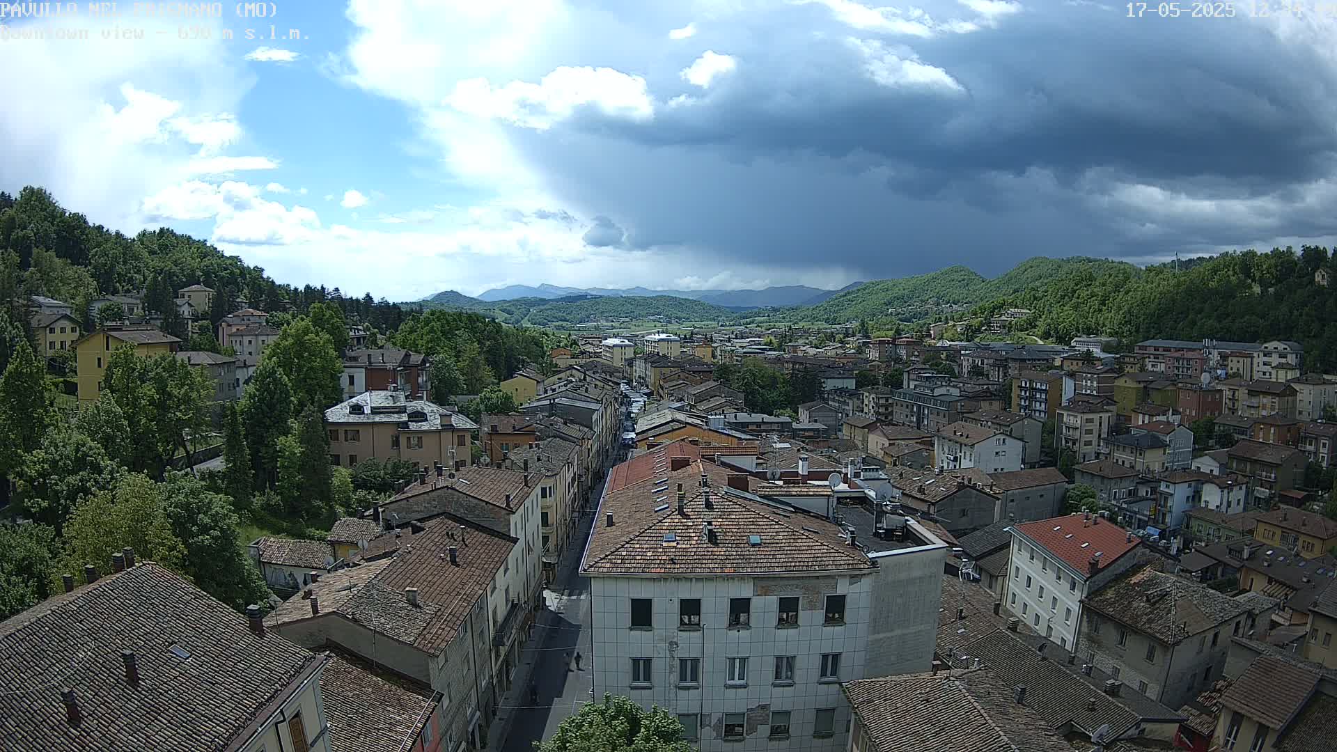 An aerial view of a town nestled in a valley, under a partly cloudy sky with dark storm clouds gathering in the distance.
