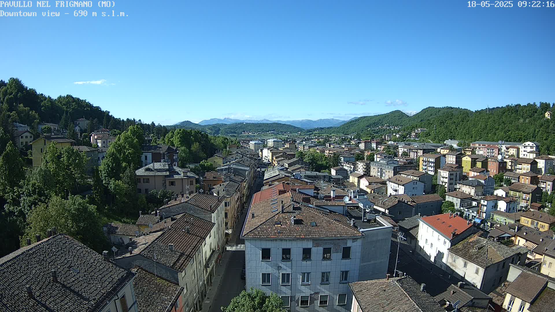 A sprawling town nestled in a valley under a clear blue sky, surrounded by verdant hills and mountains in the distance.