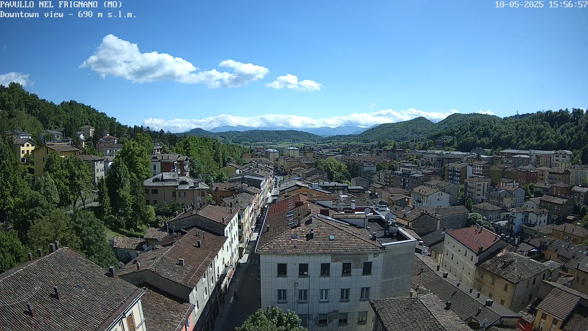 A high-angle view of a town nestled in a valley under a mostly sunny sky with some scattered clouds and distant mountains.