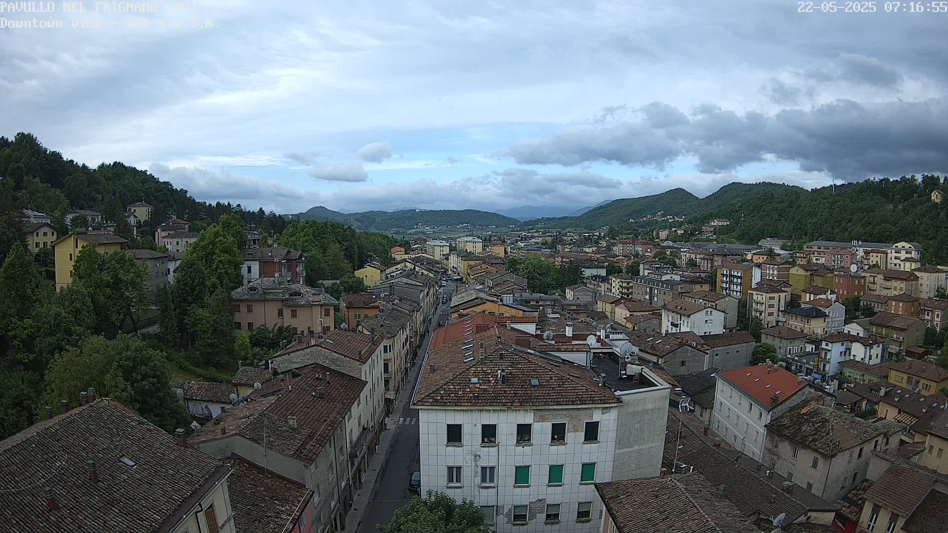 A partly cloudy day reveals a view of a town nestled in a valley, surrounded by green hills.