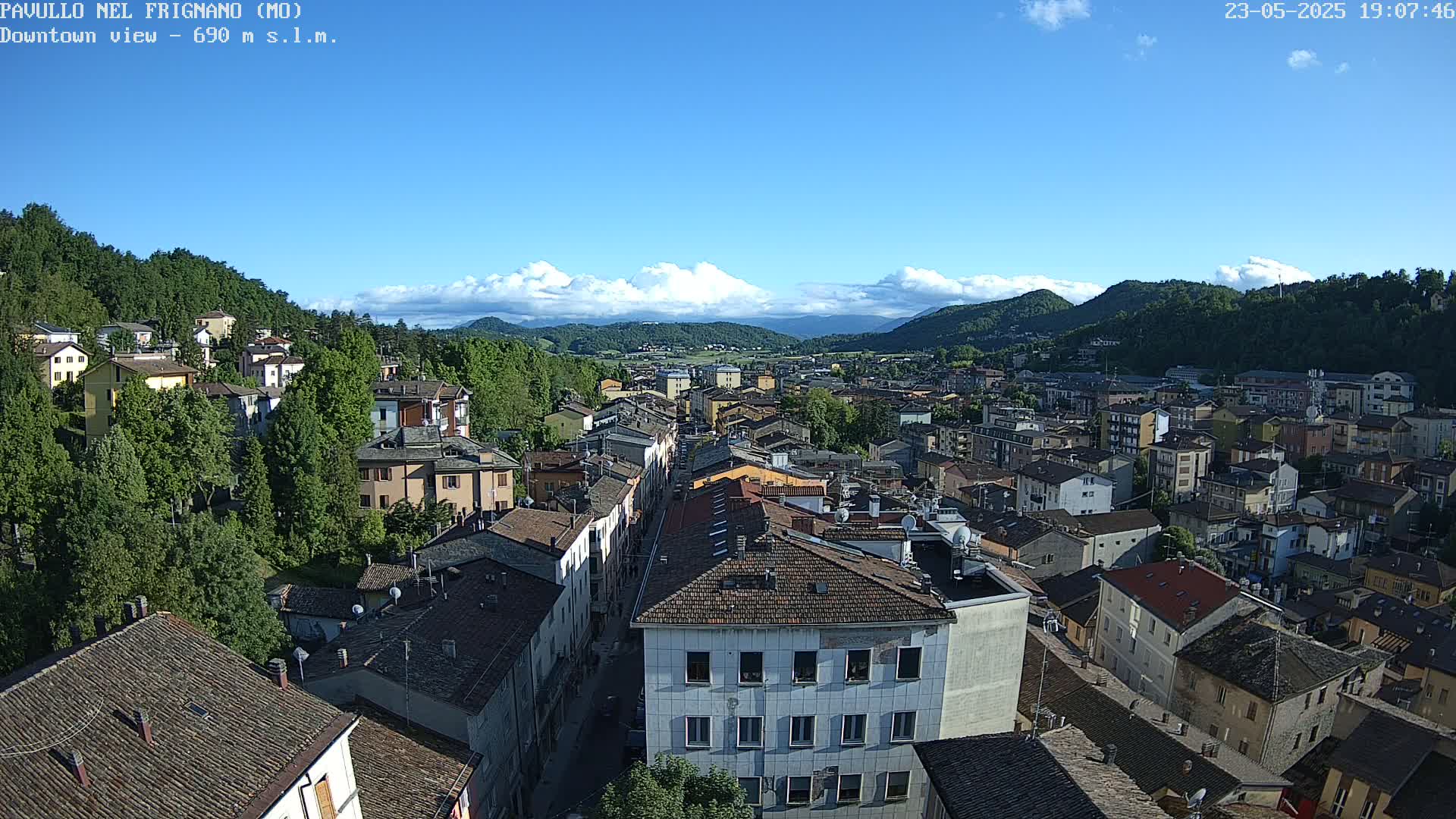 A mostly sunny day reveals a high-angle view of a town nestled in a valley surrounded by hills and mountains, with some cumulus clouds in the sky.