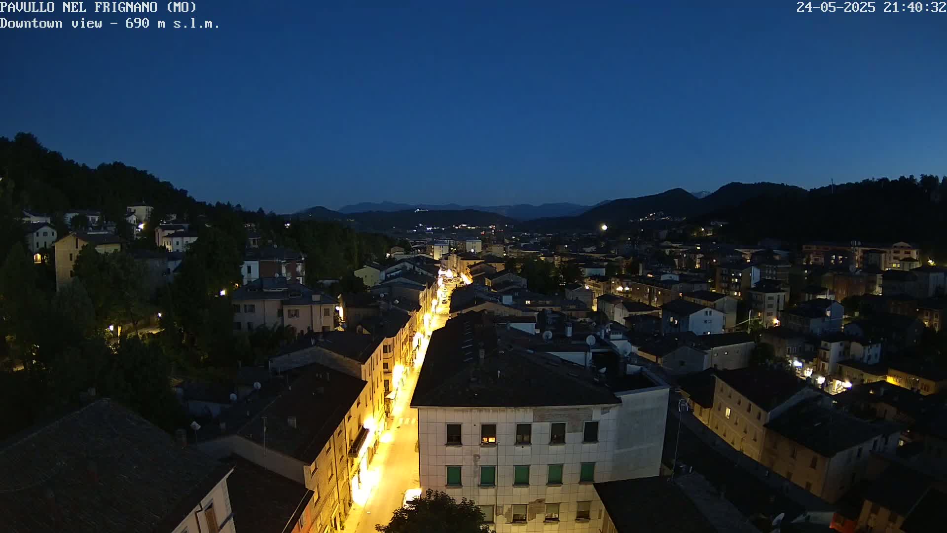 An aerial nighttime view of a town nestled in a valley, with a long street brightly lit, under a clear, dark blue twilight sky.